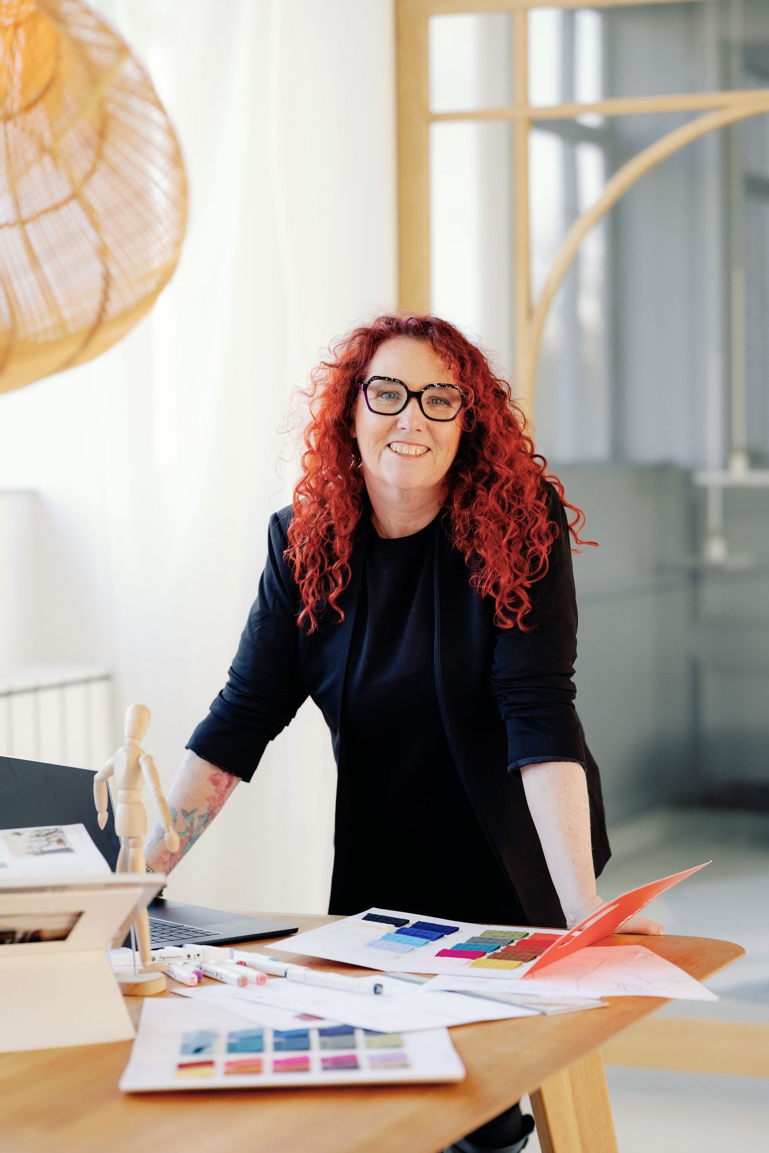 Person with red hair works at desk reviewing colorful design materials in bright office space.