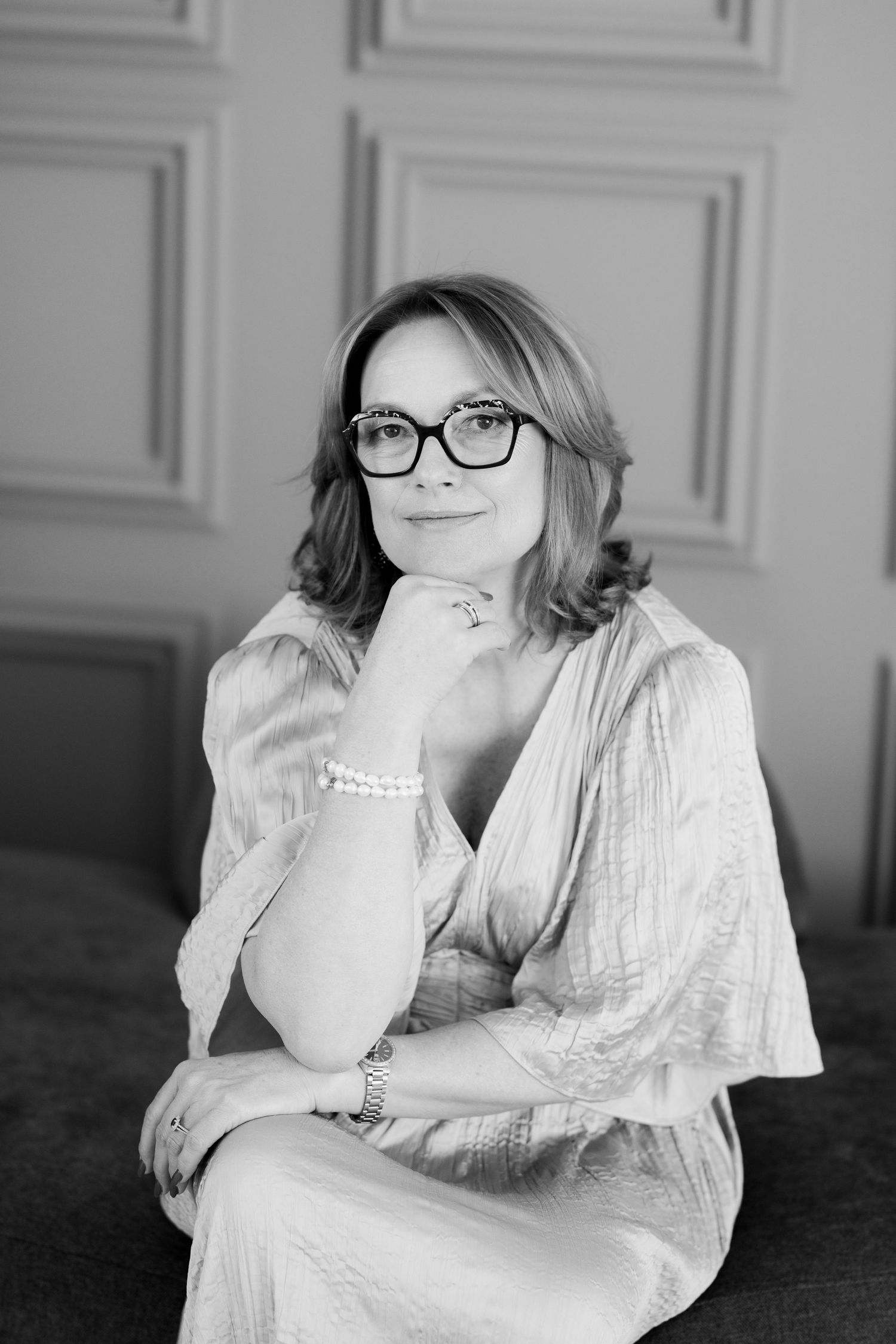 Black and white portrait of a person wearing glasses and a light colored top in an elegant indoor setting.