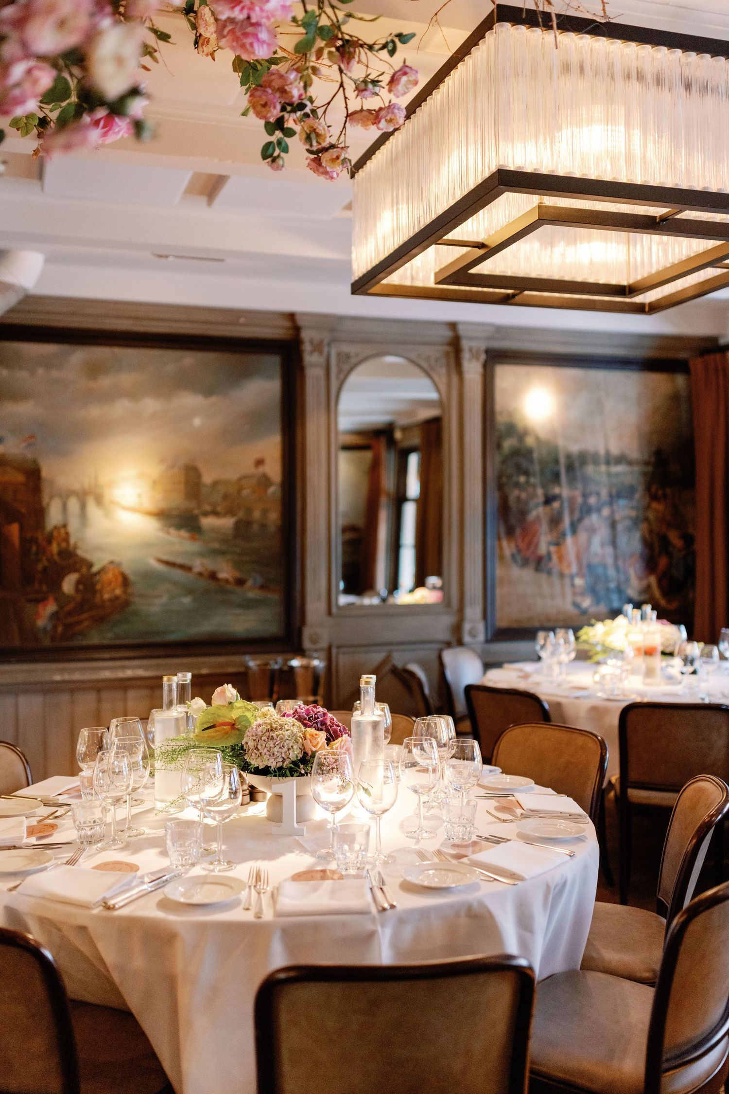 Elegant dining room setup with round tables covered in white linens, crystal glassware, and ornate wall paintings.