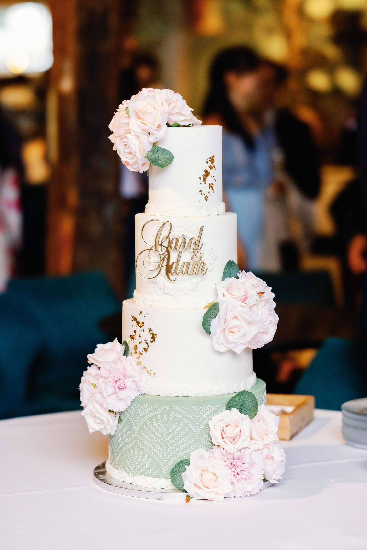 An elegant three-tiered wedding cake decorated with sage green accents and delicate pink roses sits on a white tablecloth.