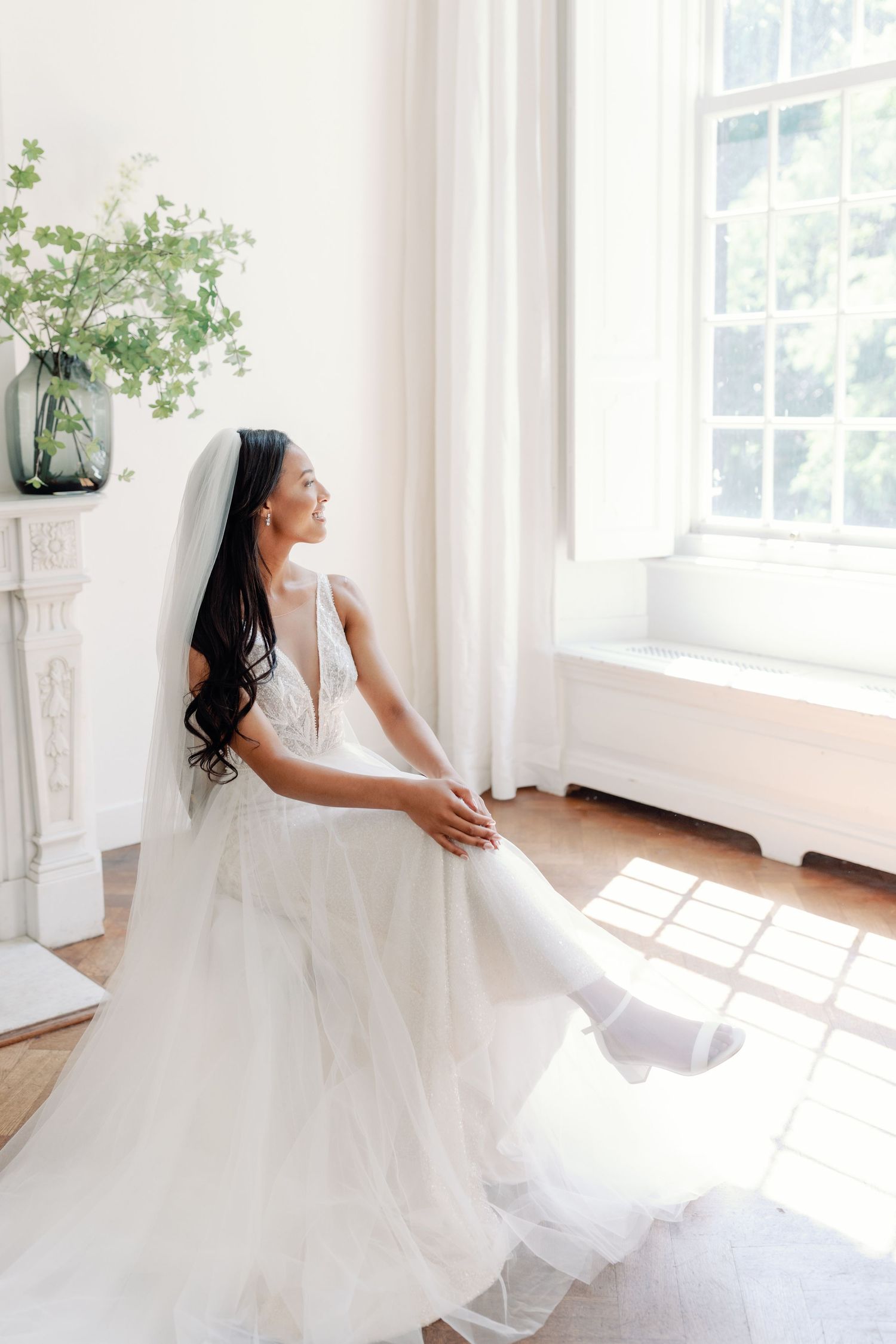 A bride in a flowing white ballgown twirls gracefully in a sunlit room with large windows.