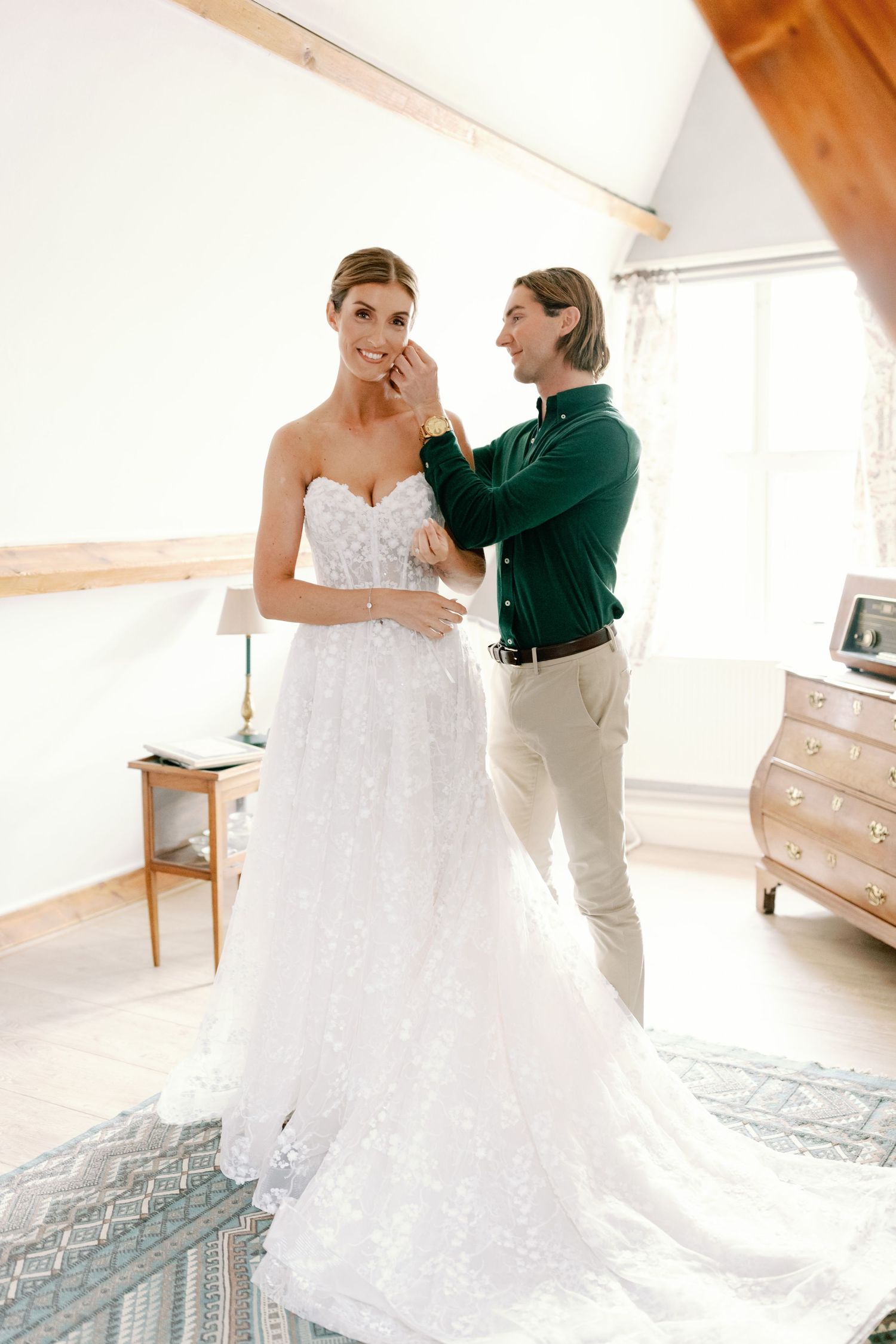 A bride in a strapless lace wedding gown stands in a sunlit room while getting ready for her special day.