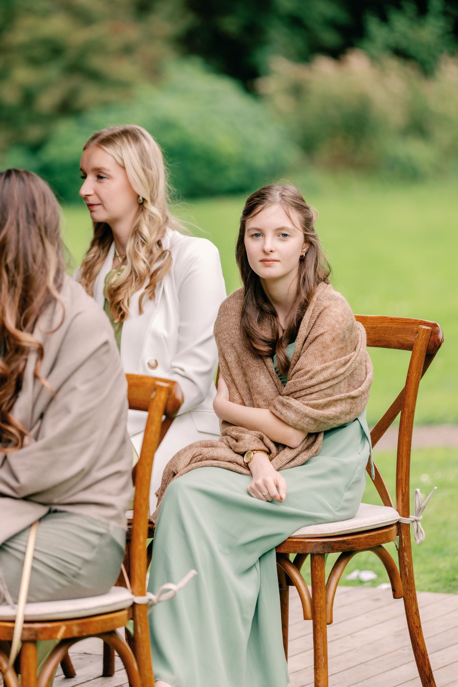 People in earth-toned dresses sit on wooden chairs on an outdoor deck with greenery in the background.