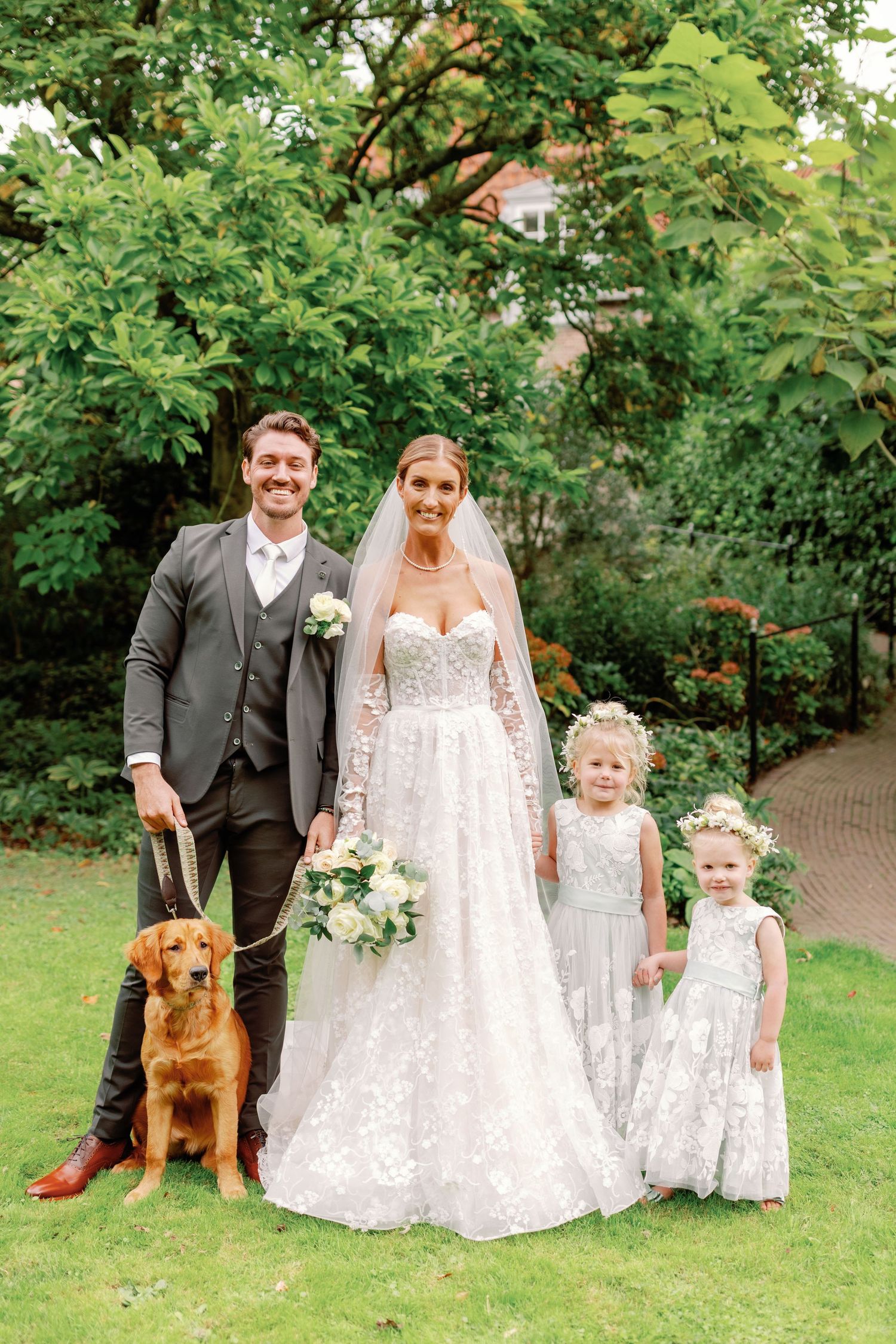 Wedding party poses on garden lawn with golden retriever dog, wearing elegant white and gray attire.