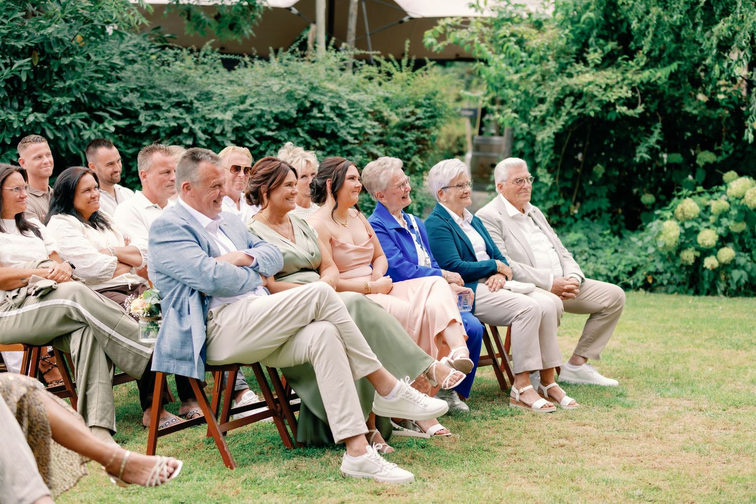 Wedding guests sit in white chairs on a lawn during an outdoor garden ceremony, arranged in multiple rows.