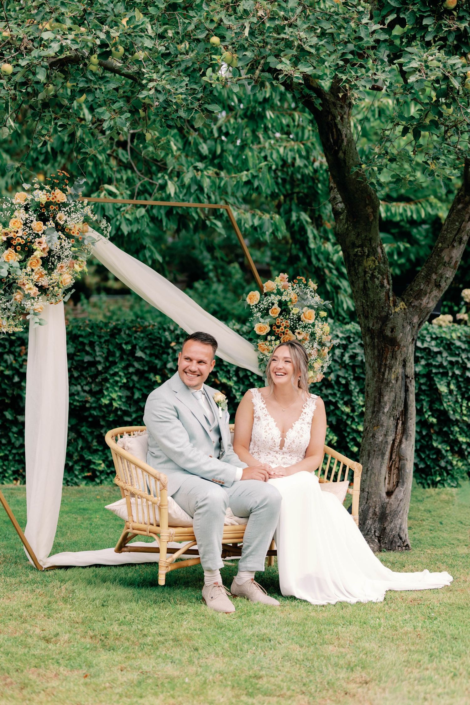 Newlyweds share a joyful moment on a rattan bench under a floral arch and tree in an outdoor garden setting.
