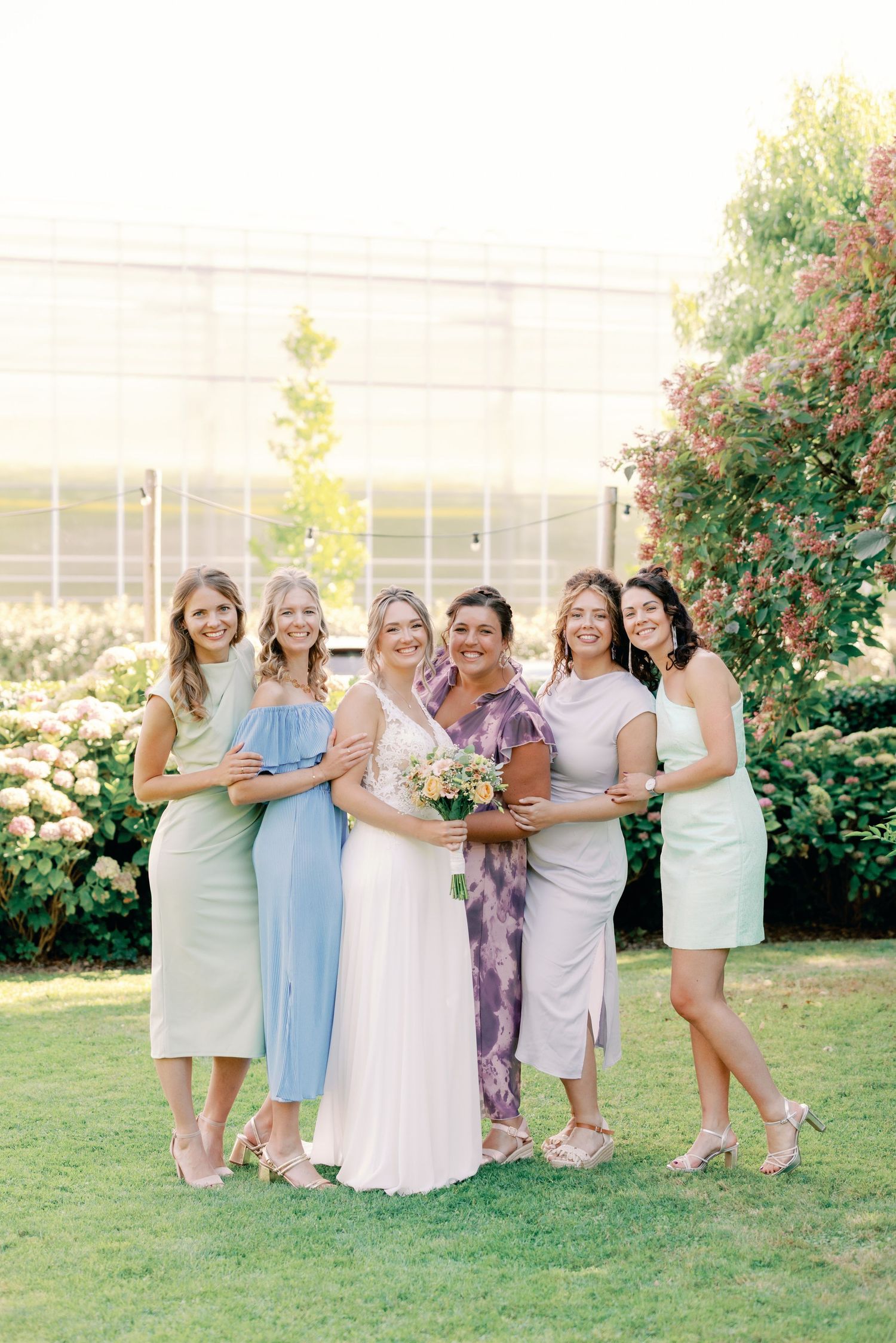 A group in pastel dresses laughs together on a sunny lawn with blooming shrubs in the background.