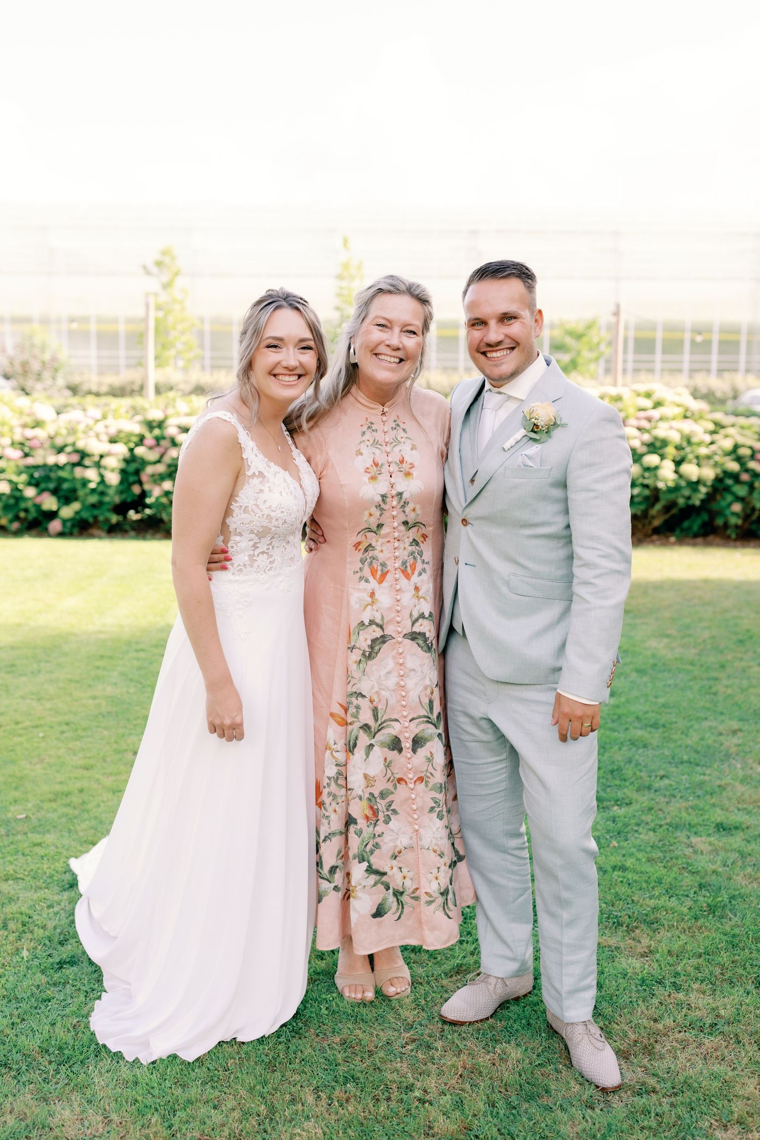 Three people pose together on a grassy lawn during a summer wedding celebration.