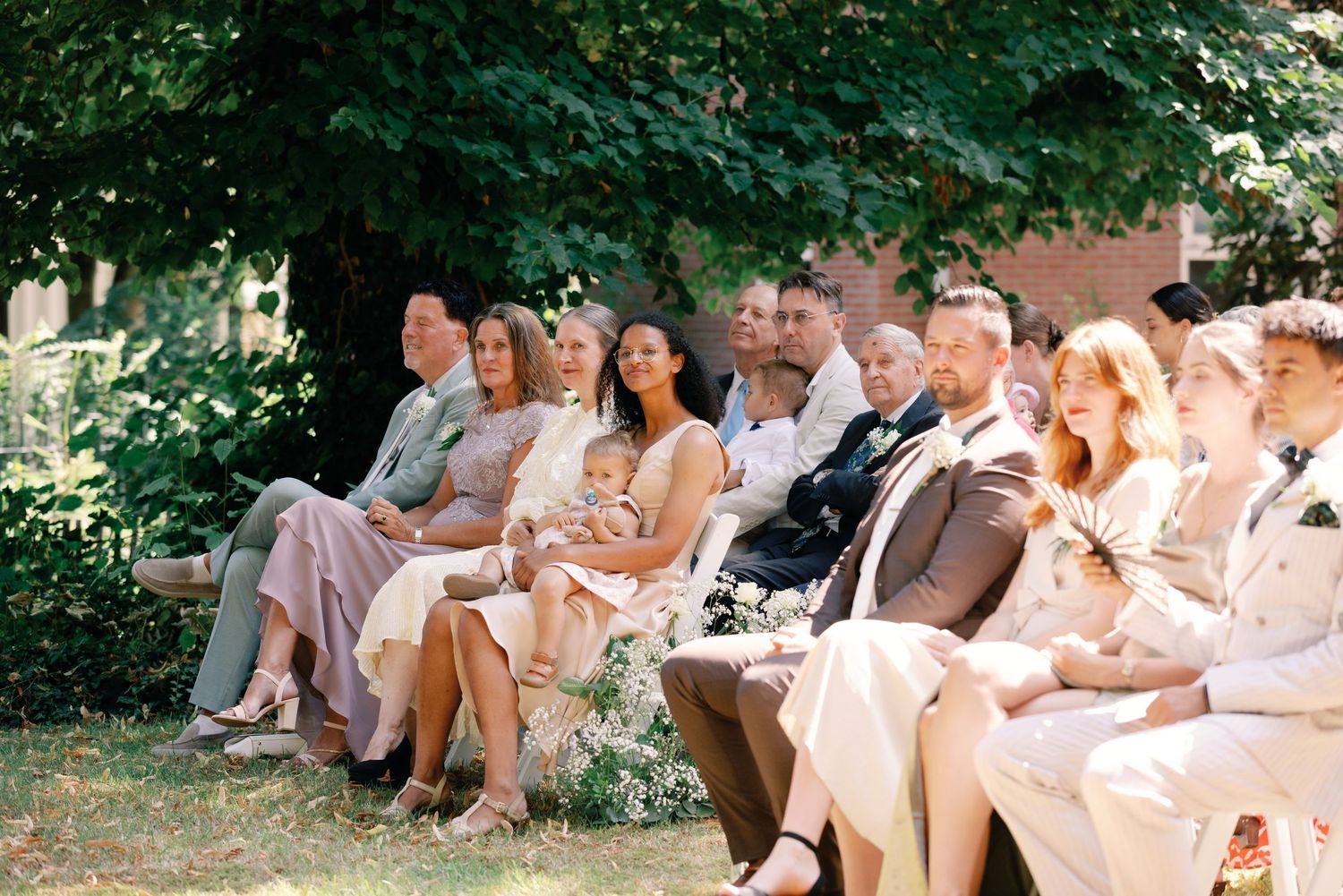 A group of people in light colored clothing sit together outdoors on a sunny day during a casual gathering.