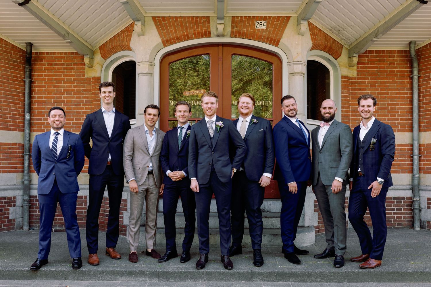 A group of groomsmen in navy suits pose playfully in a line against a brick building exterior.