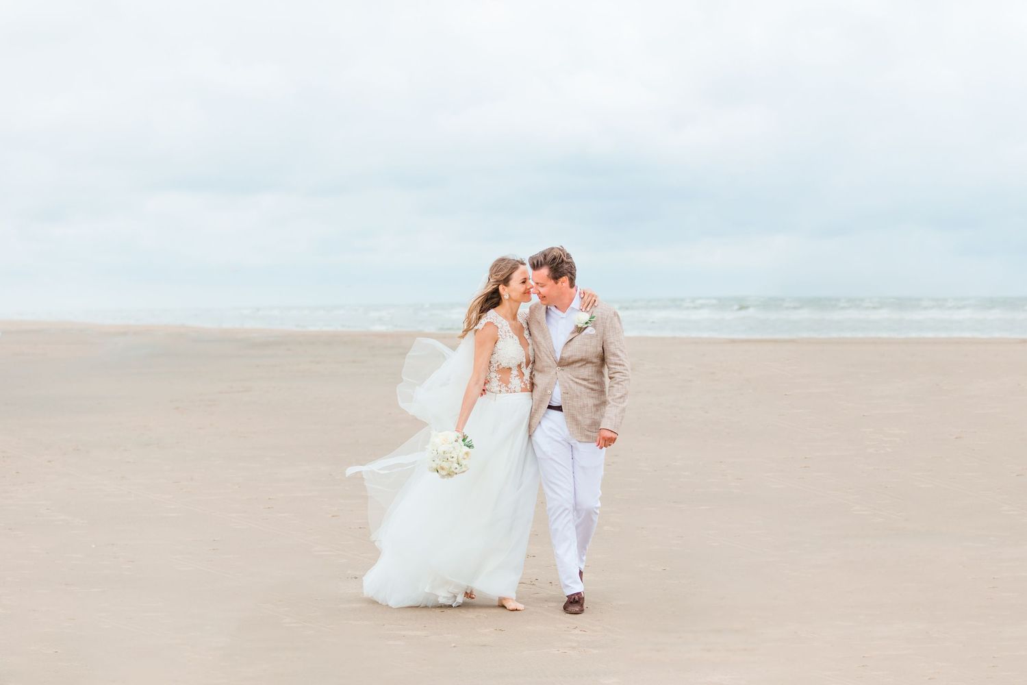 A romantic beach wedding couple walking together on sandy shore with cloudy sky in the background.