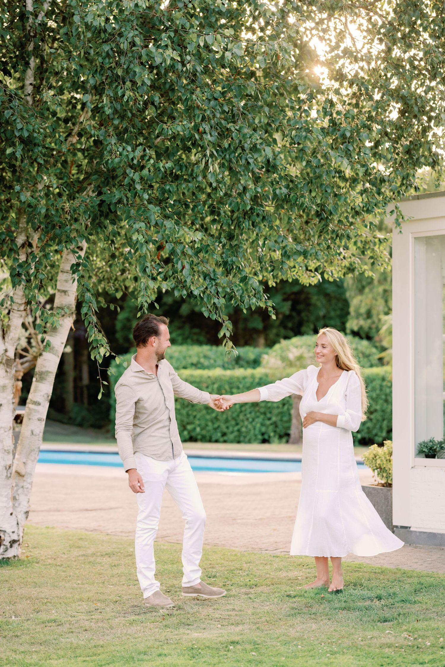 Couple dancing and twirling in white outfits against a backdrop of green trees and a pool.