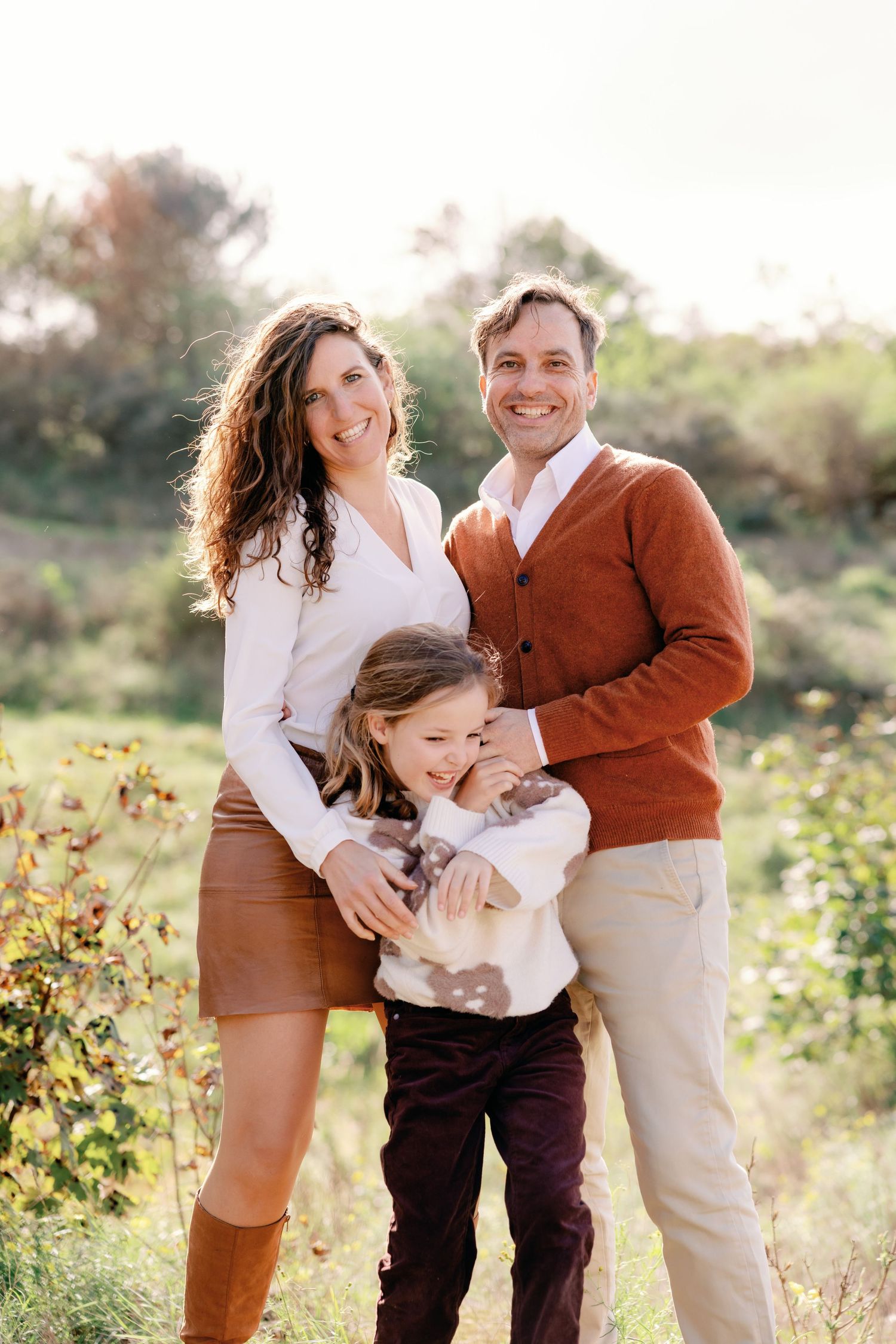 A happy family of three poses outdoors in autumn, wearing warm earth tones, with a child hugging her parents.