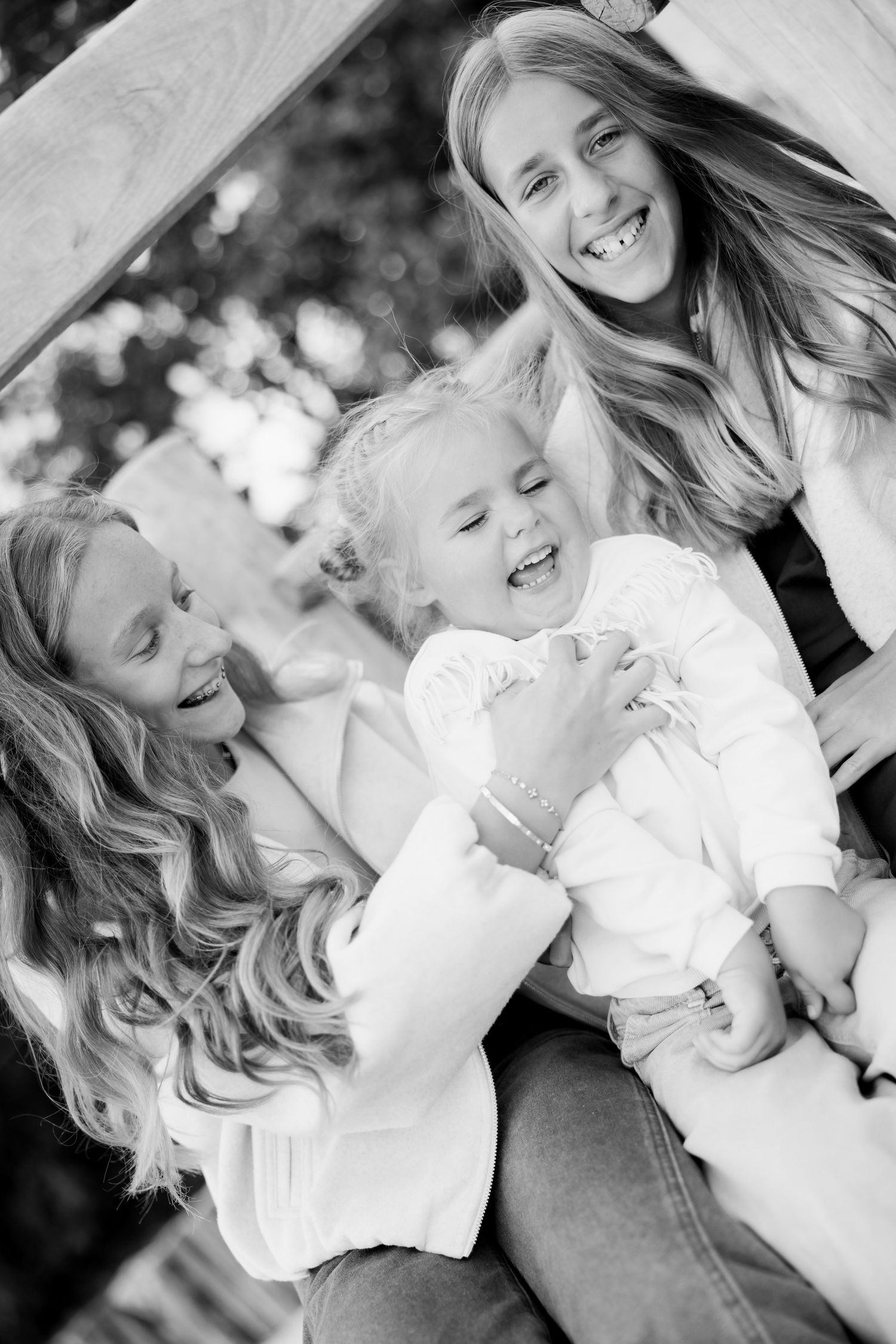 Black and white photo of two smiling girls and a laughing toddler cuddled together outdoors on a wooden bench in a joyful family moment.