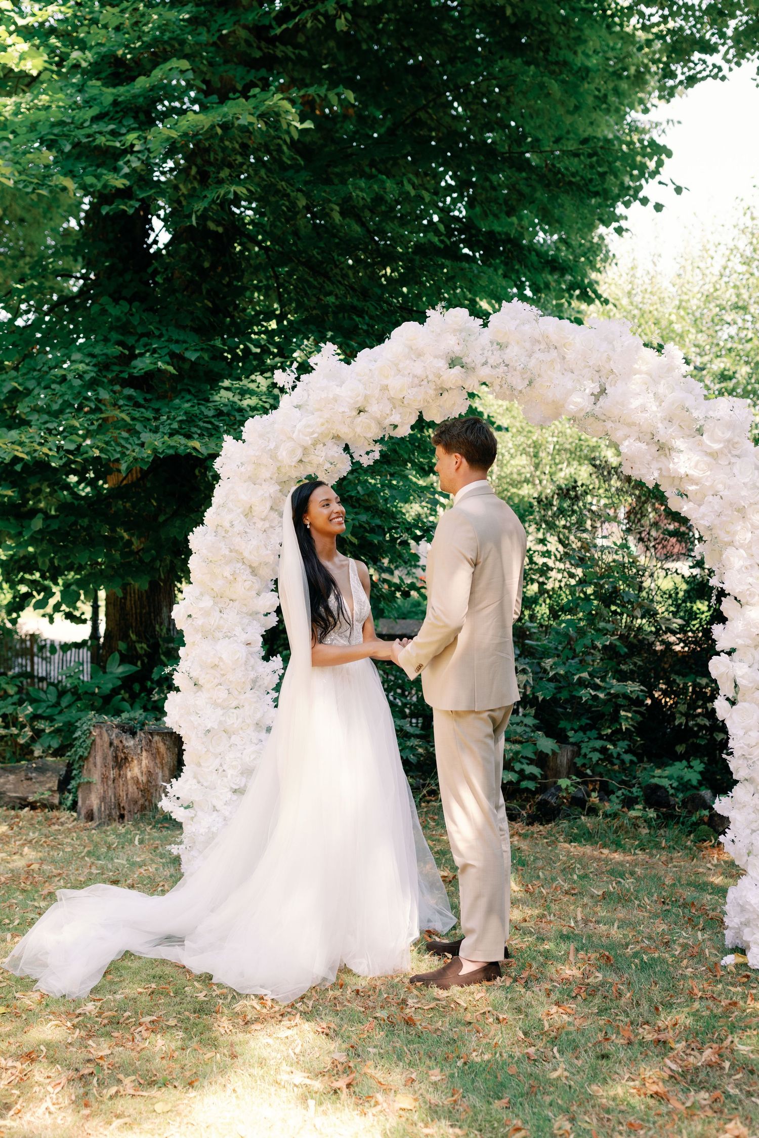 A beautiful outdoor wedding ceremony setup with a white floral arch and couple exchanging vows in a garden setting.
