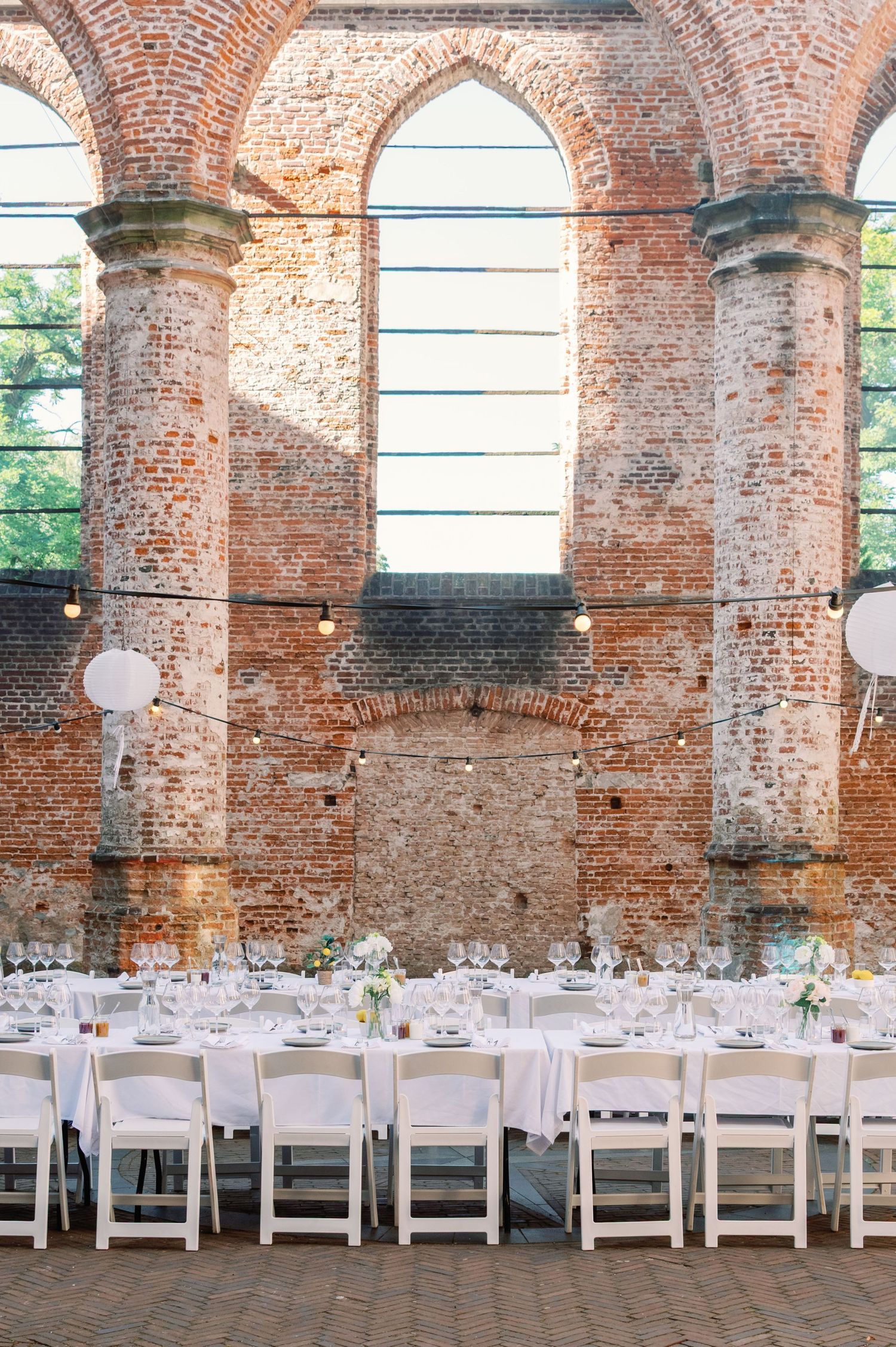 Long banquet tables with white linens set for a wedding reception inside historic brick ruins with arched windows.