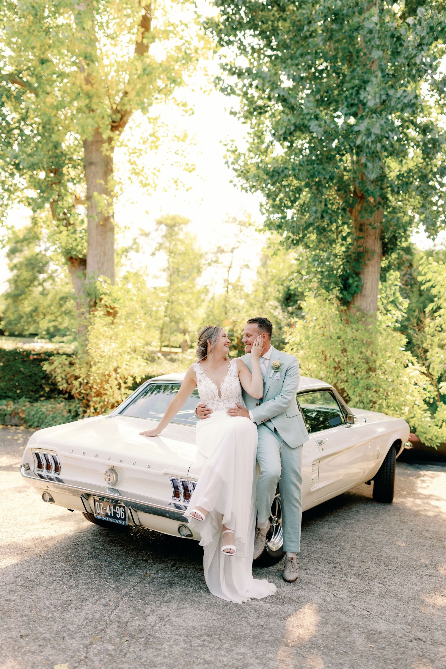 A classic white vintage car parked under green trees creates an elegant backdrop for a romantic outdoor scene.