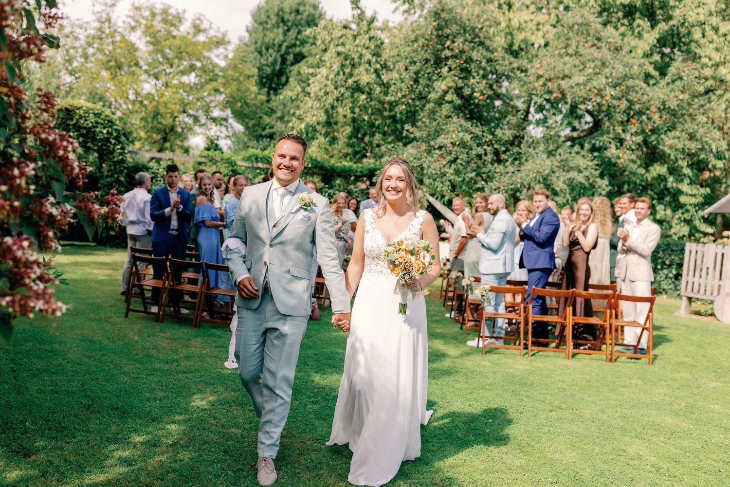 Outdoor wedding ceremony in a garden setting with wooden chairs and guests gathered on a sunny day.