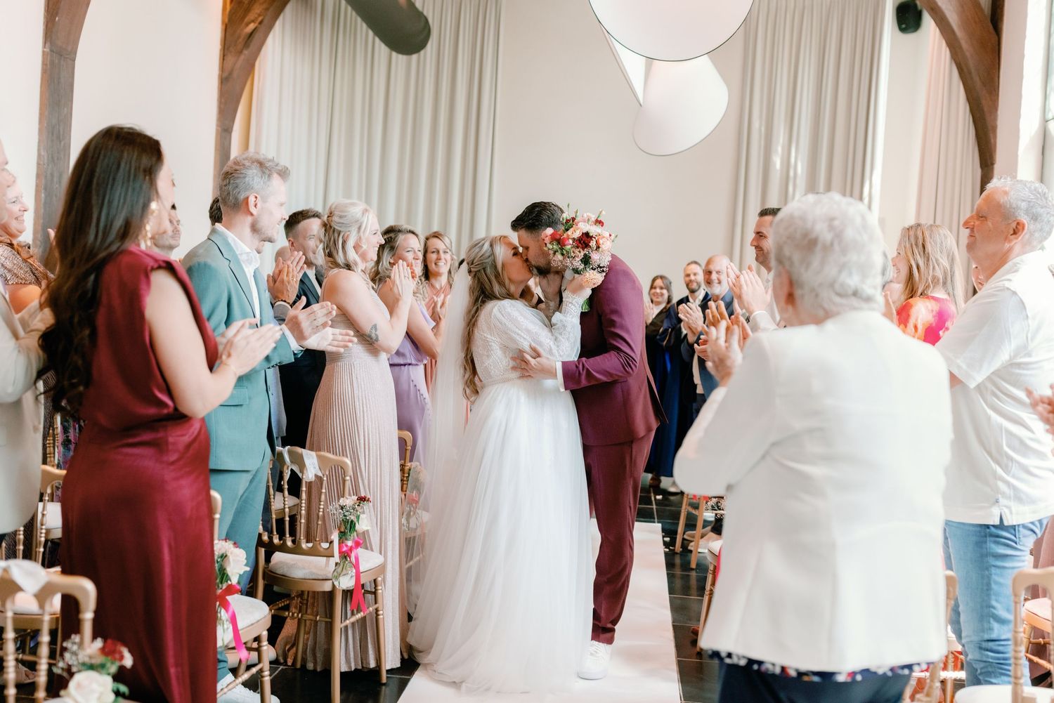A wedding party celebrates with guests in a bright indoor venue with white curtains and decor.