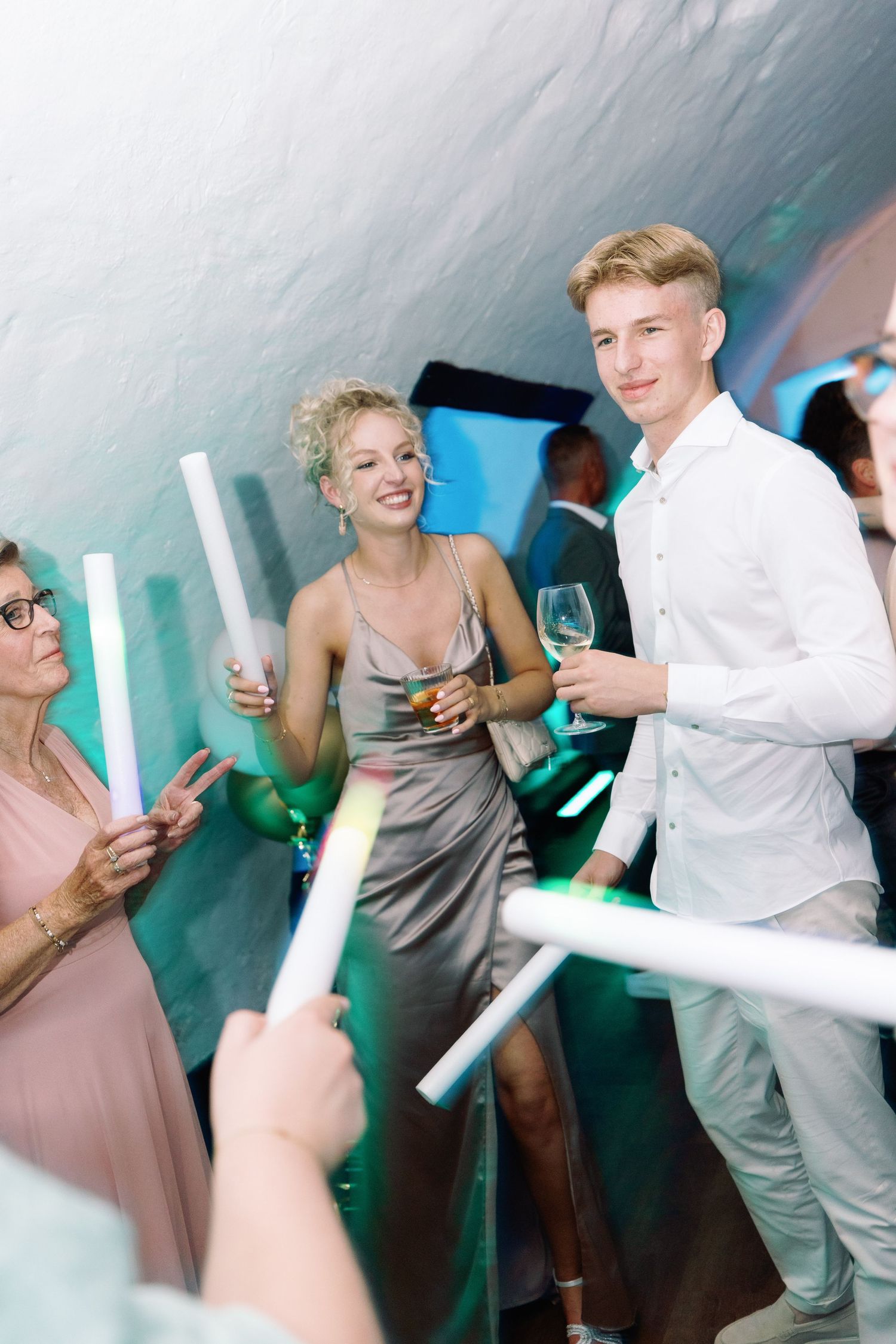 Wedding guests dancing and socializing at a reception with turquoise lighting and festive atmosphere.