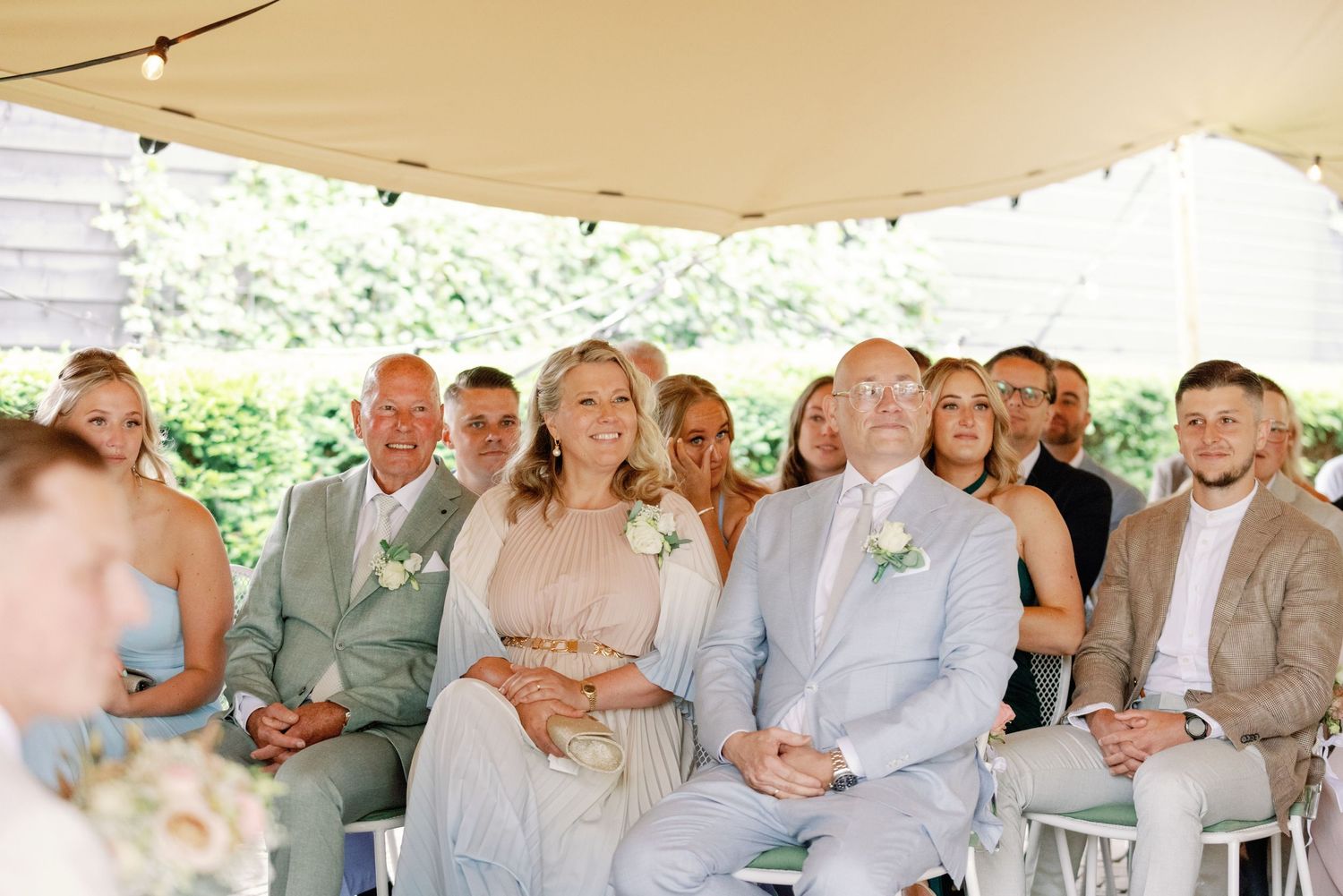 A row of wedding guests under a white tent watching the celebration with string lights overhead.