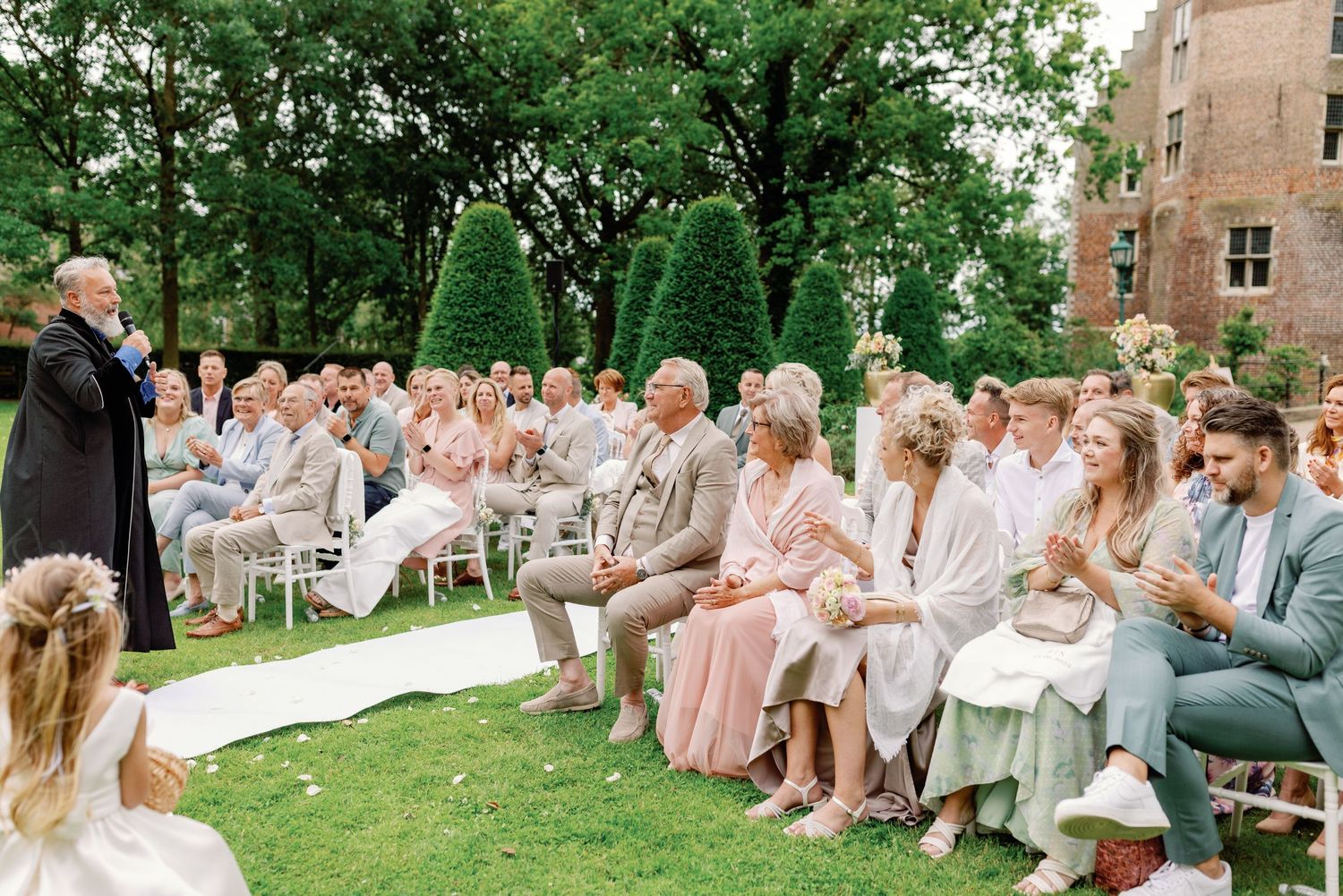Wedding guests seated on white chairs during an outdoor garden ceremony with lush greenery and trees.