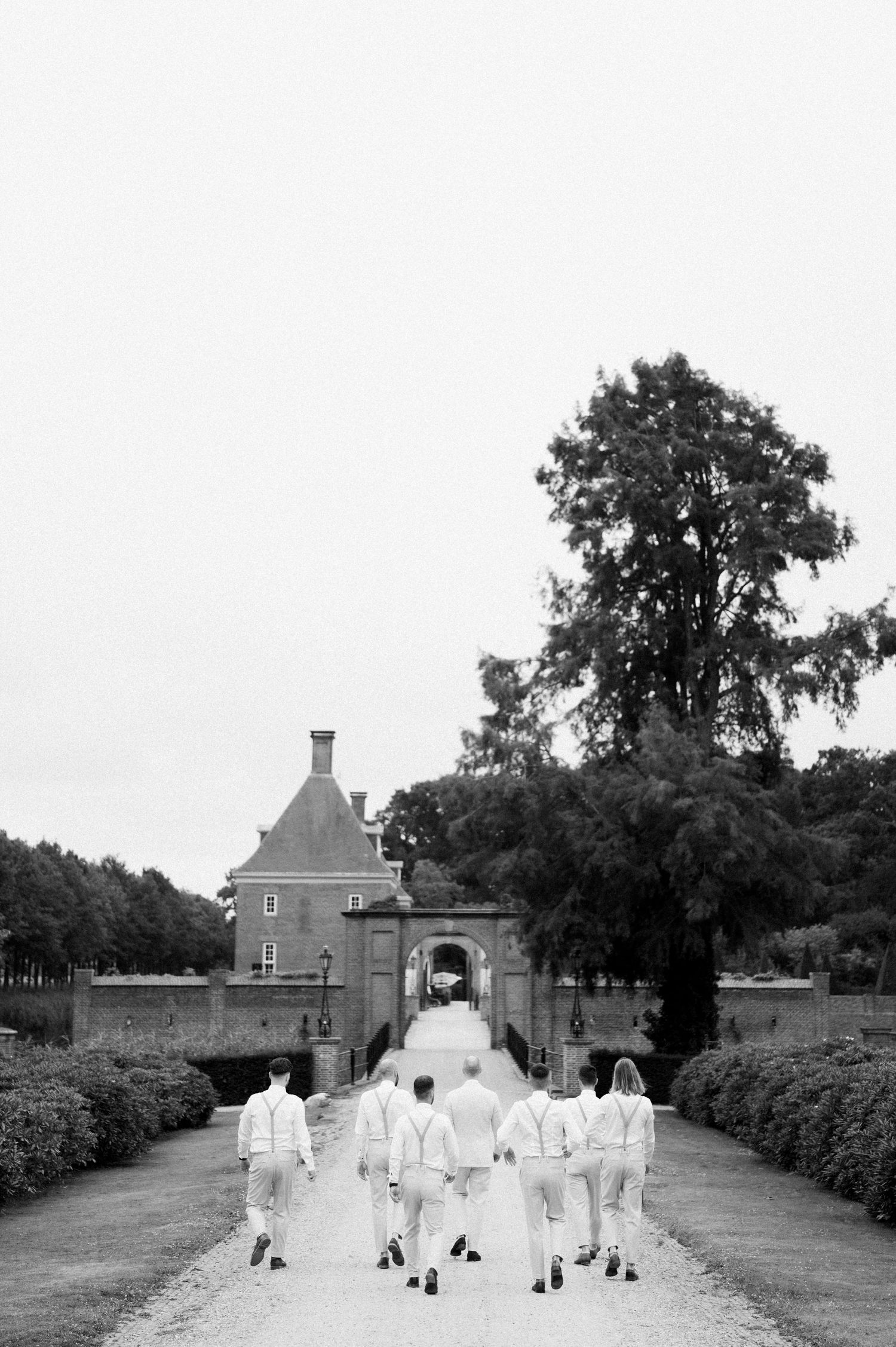 A black and white photo shows a group in white robes walking in a line towards a brick building with trees.