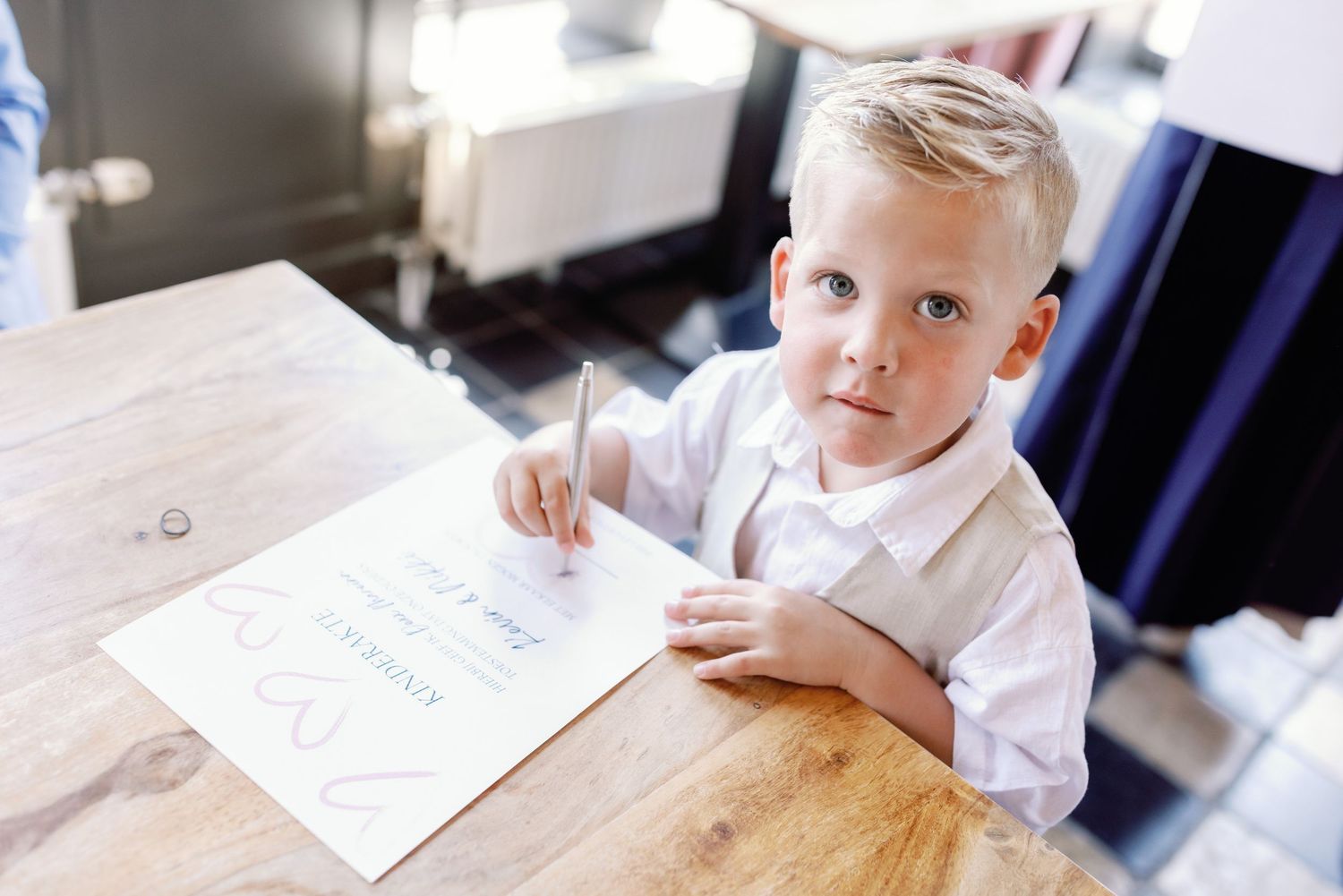 Young blonde boy in white shirt writing on paper at wooden table, looking up at camera.
