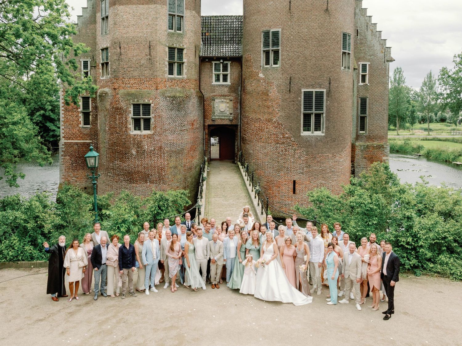 Large wedding party poses in front of historic brick castle building during outdoor summer celebration.