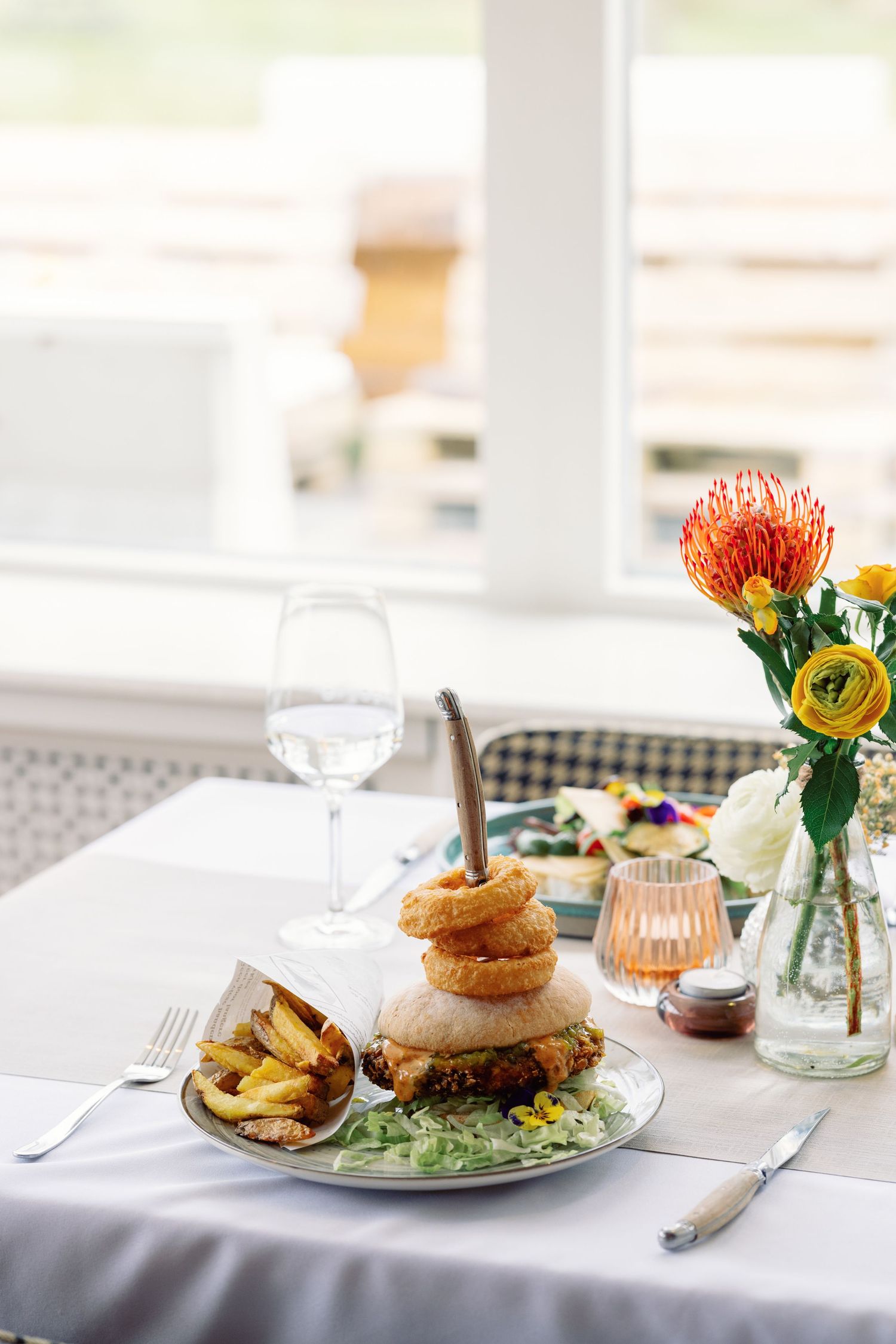 Gourmet burger with onion rings and fries served on white plate with floral centerpiece and wine glass in bright restaurant.