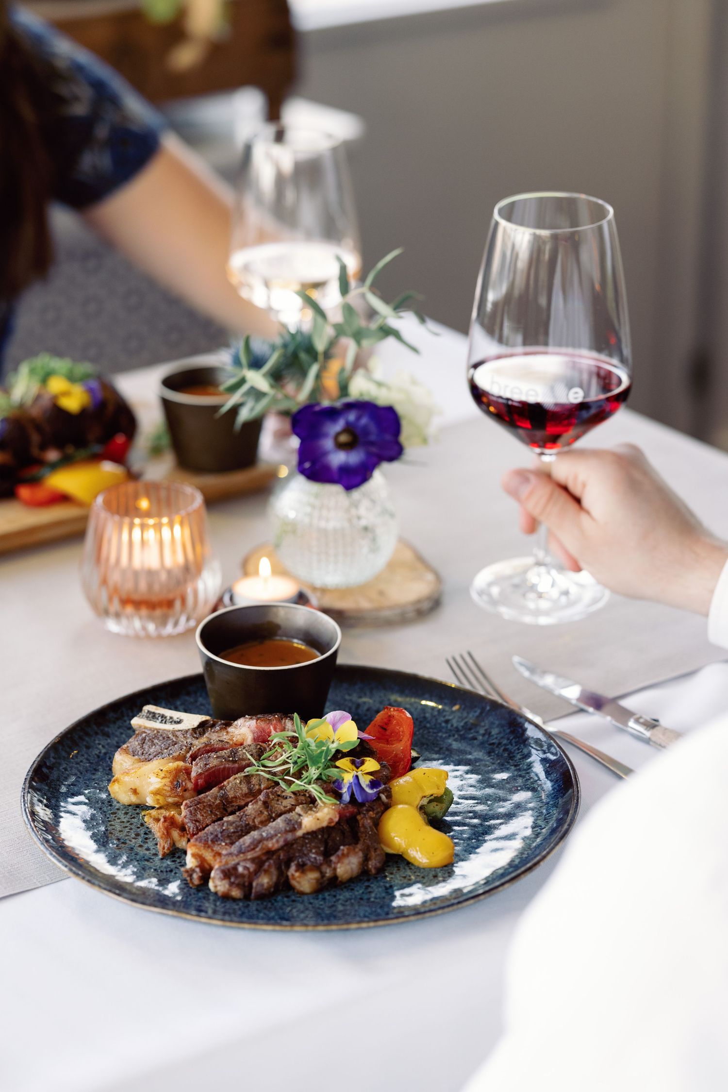 Elegant plated dishes with grilled meats and colorful garnishes served on dark plates against a white tablecloth.