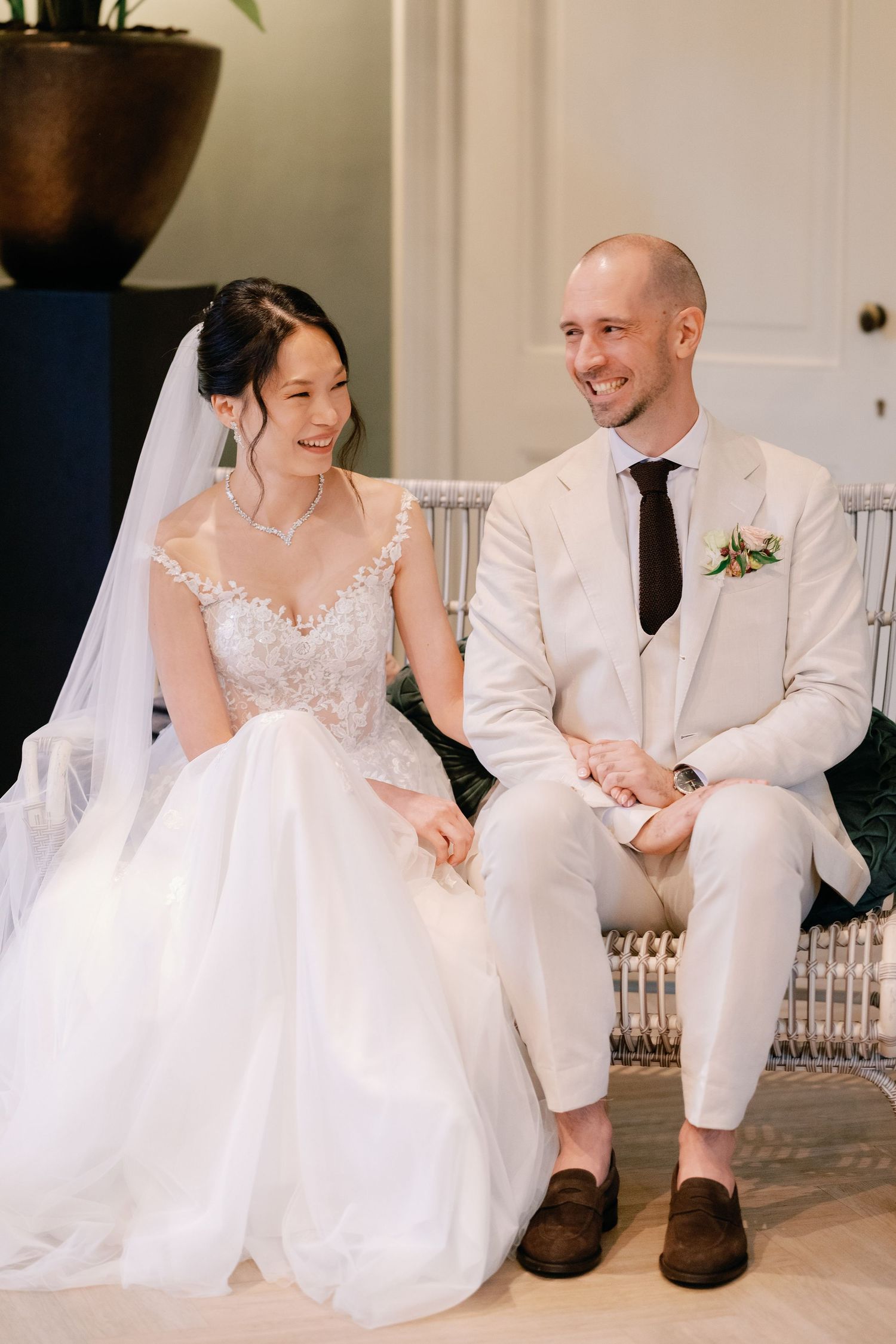 A couple in wedding attire sits together on white steps, sharing joyful moments during their special day.
