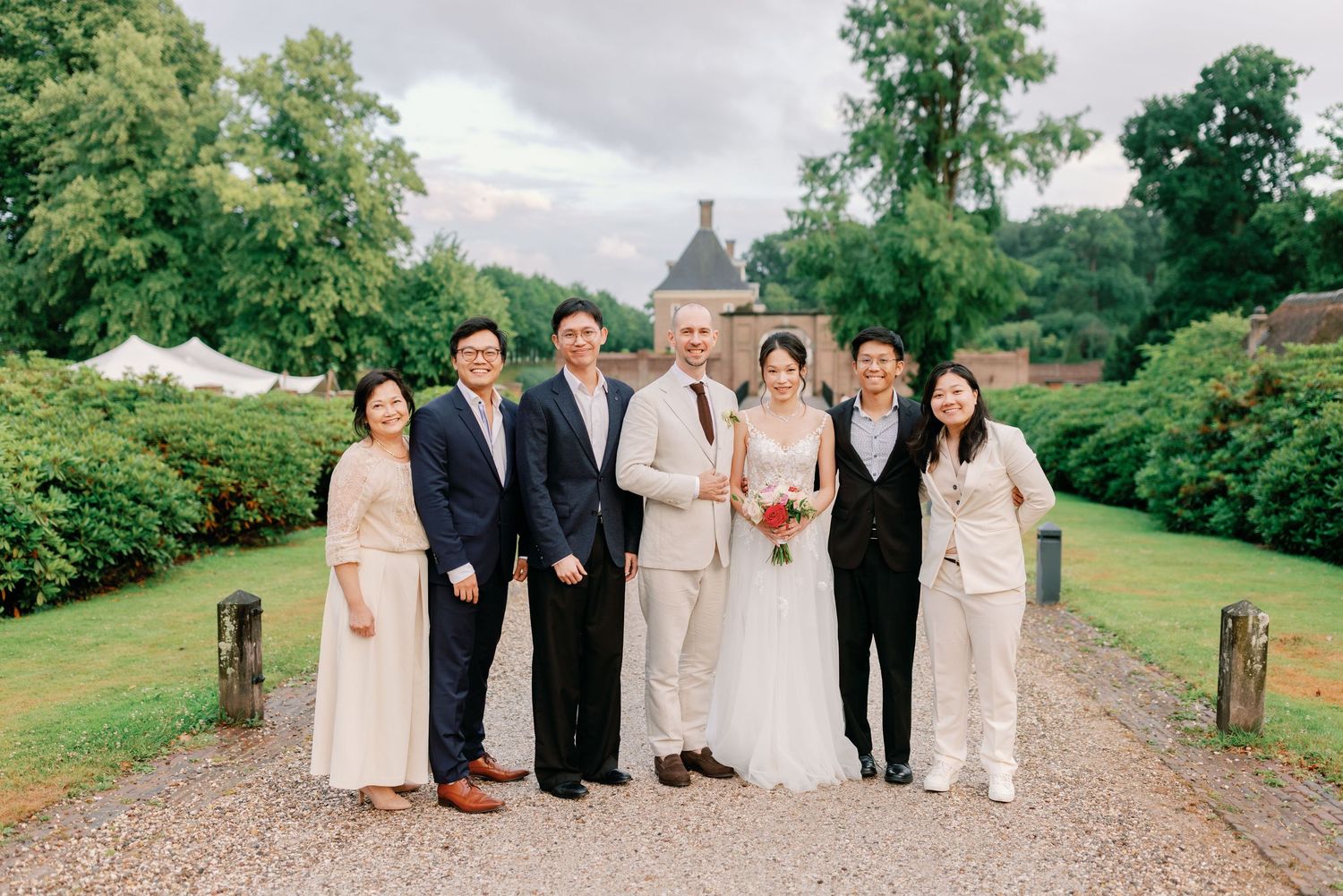 Wedding party members pose together on a gravel path surrounded by lush green trees and gardens.