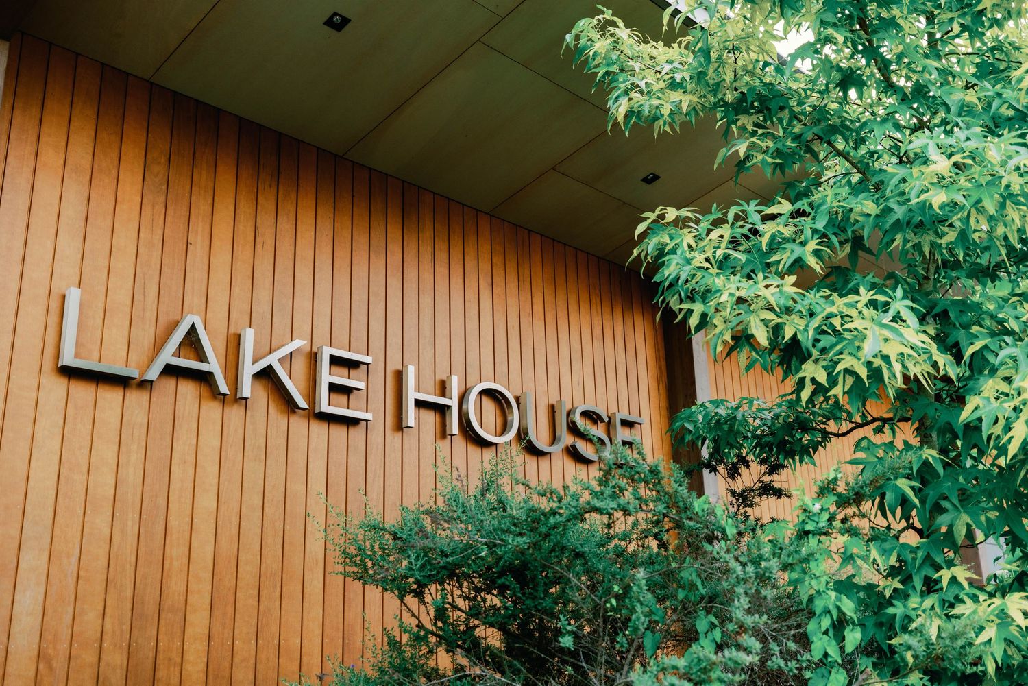 A rustic brown wooden lodge or lake house exterior with green trees visible against the natural siding.