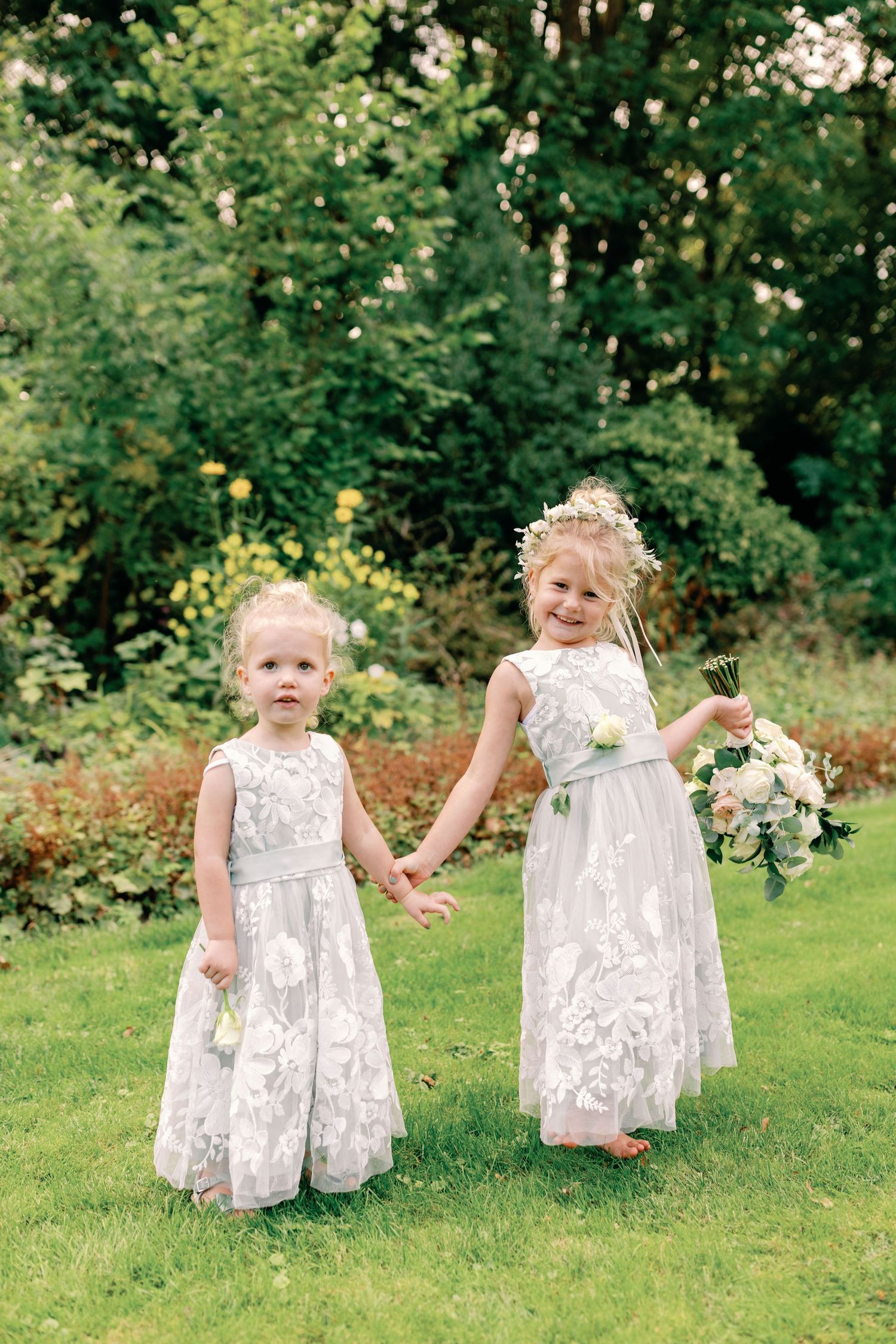 Two small children in white lace dresses play together on a green lawn surrounded by trees and foliage.