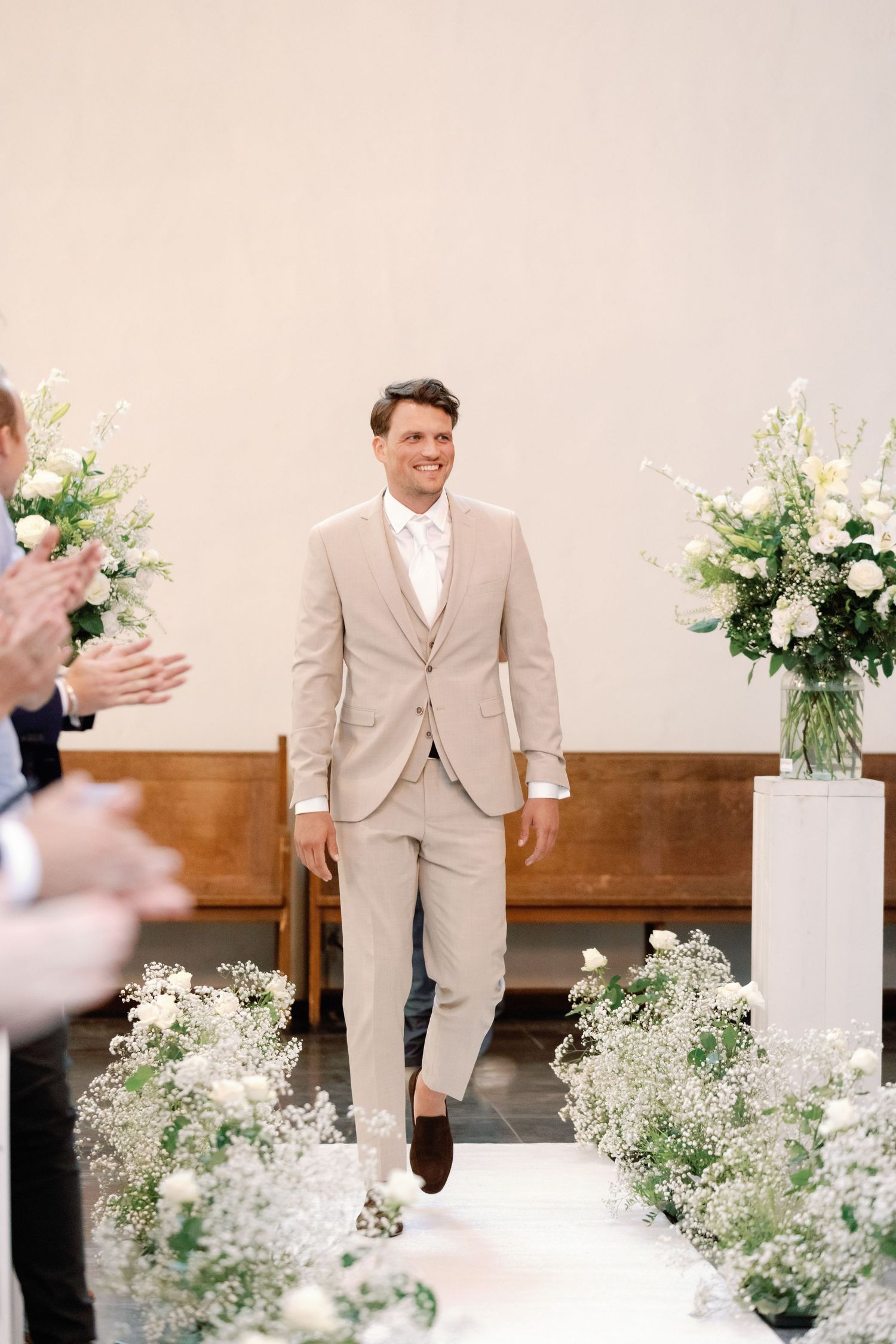 A groom in a beige suit walks down an aisle decorated with white floral arrangements and baby's breath.