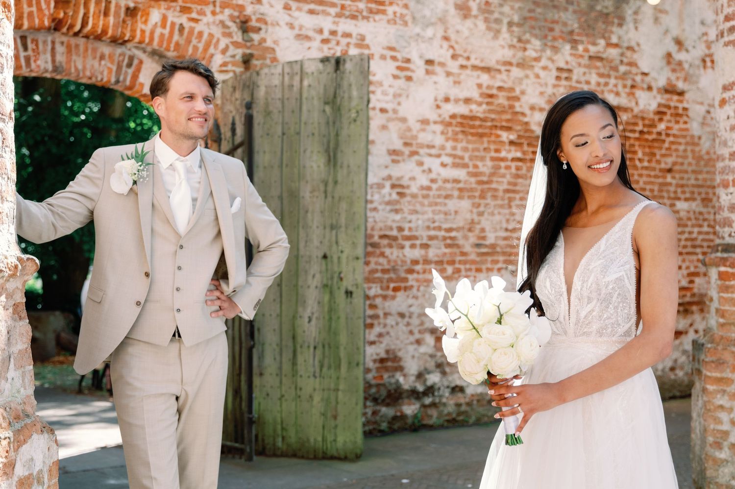 Wedding party poses in front of rustic brick wall and wooden door wearing light beige suits and white dresses.