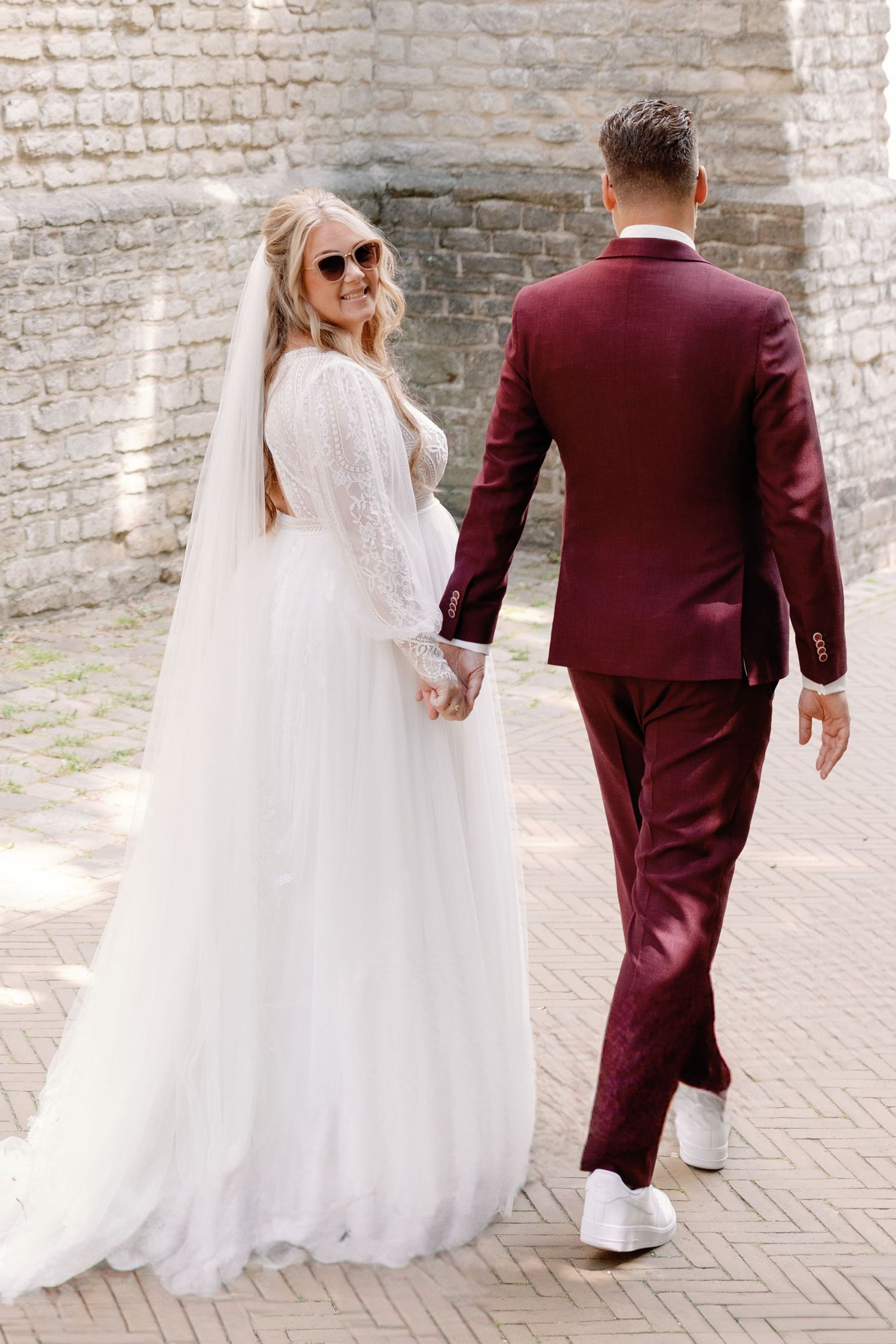 A couple in burgundy suit and white wedding dress walk together against a rustic brick wall backdrop.