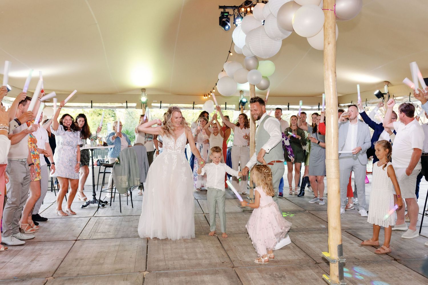 Wedding guests dance and celebrate under a white tent decorated with balloons at an outdoor reception party.