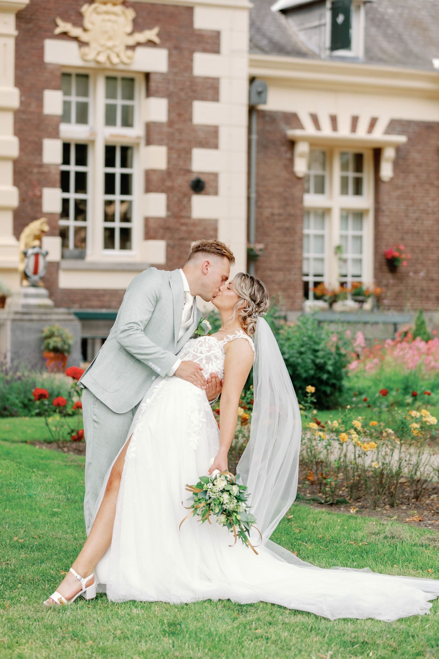 Wedding couple poses romantically on manicured lawn in front of brick building with flowers.