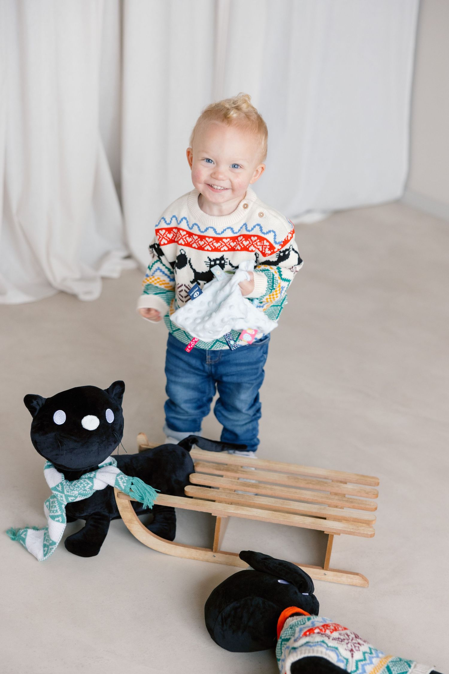 Toddler in a festive sweater playing with a black stuffed cat toy on wooden slats.