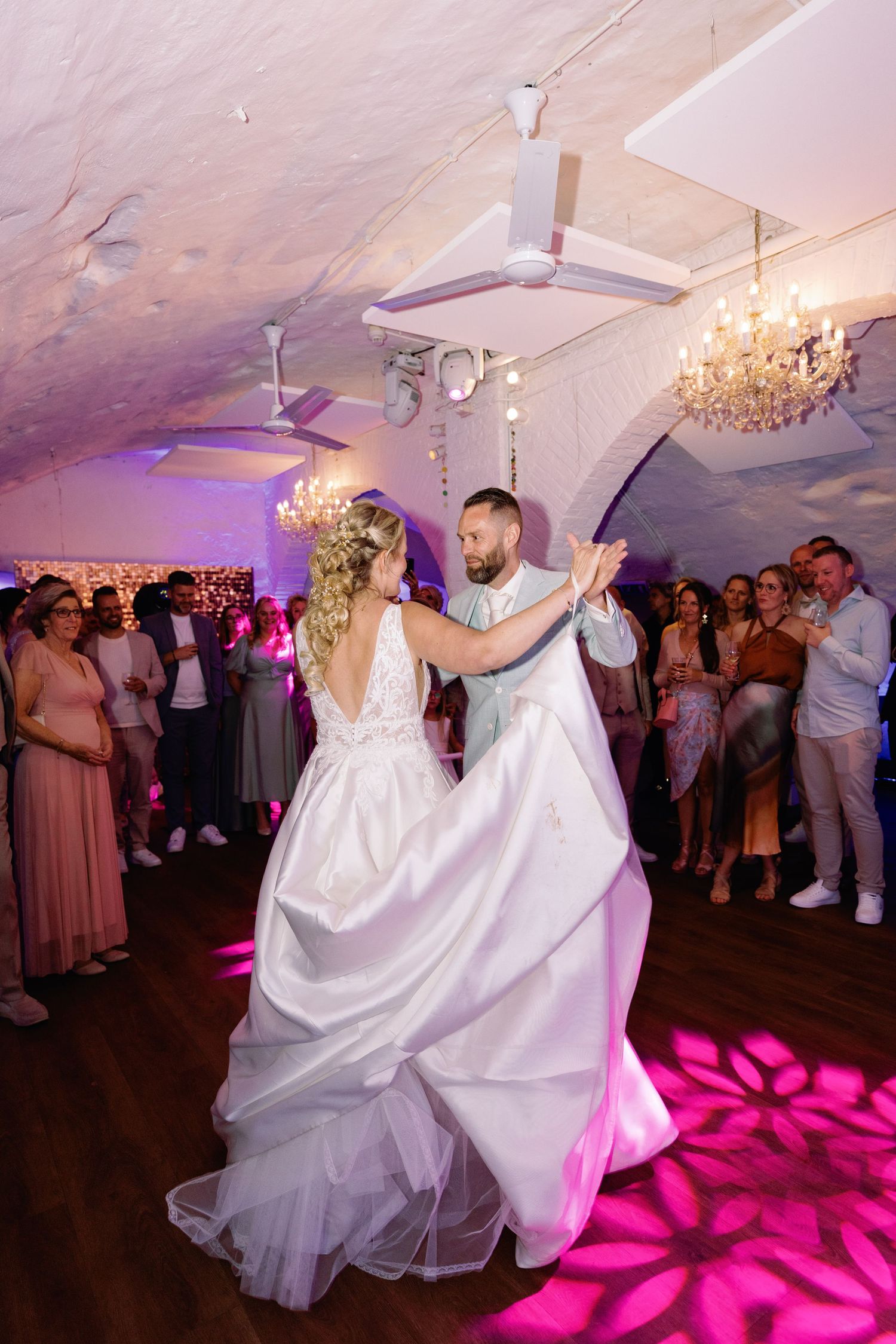 Couple performs energetic dance moves on wedding reception dance floor illuminated with vibrant pink floral lighting patterns.