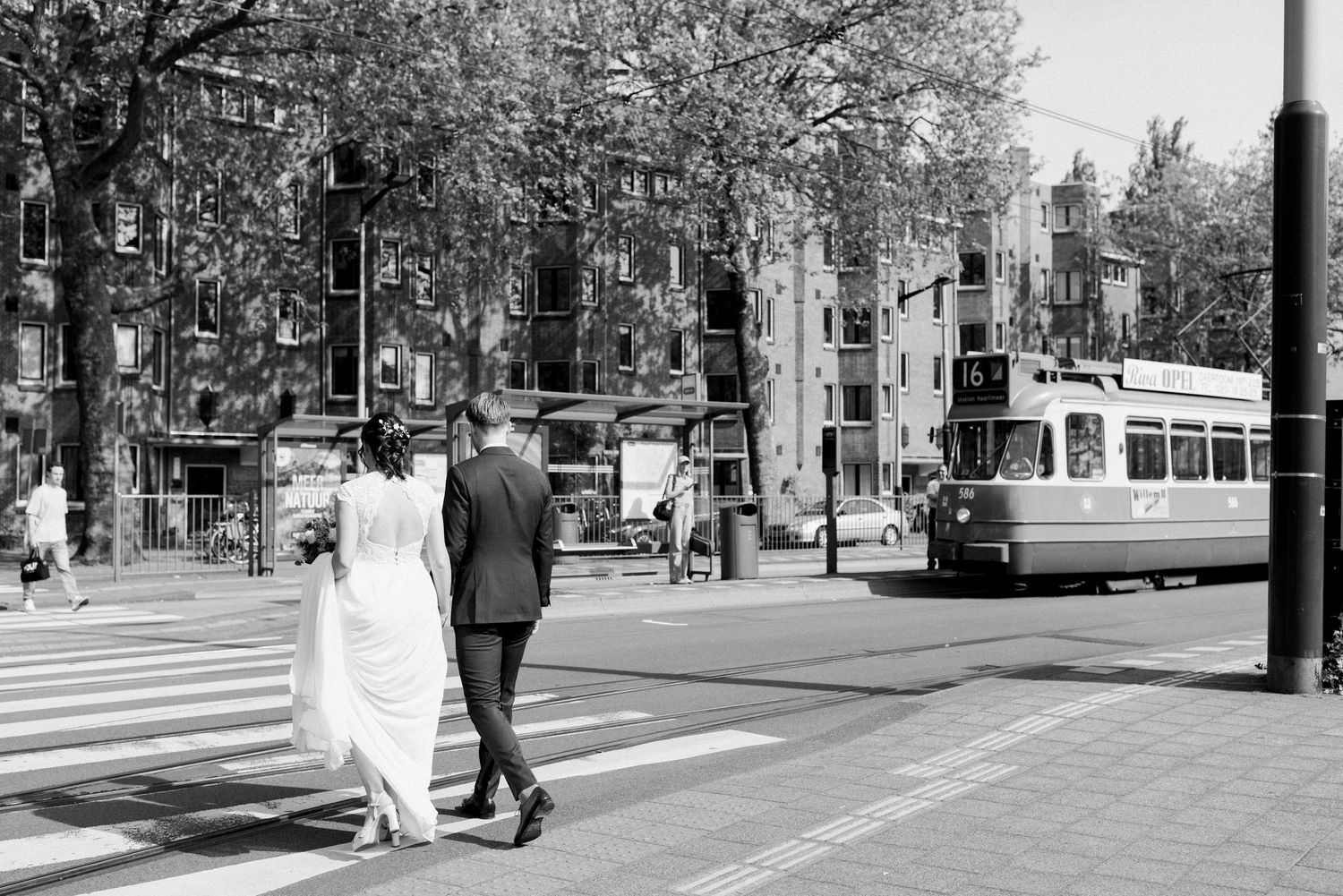 Black and white street scene showing pedestrians walking along a tree-lined urban sidewalk on a sunny day.