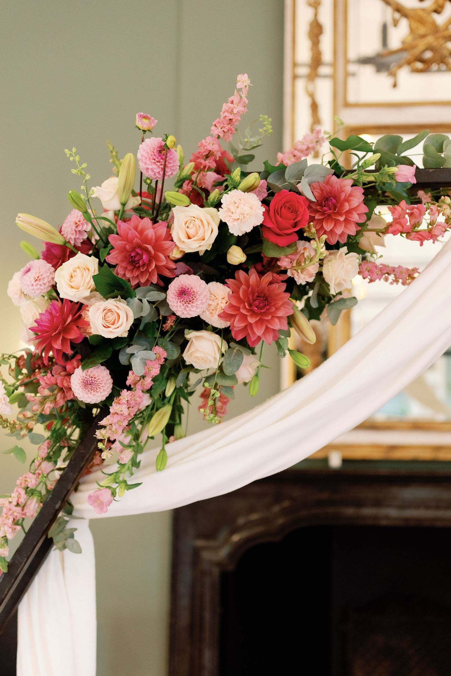 Elegant floral arrangement with pink dahlias, roses, and greenery adorning white fabric draped over a fireplace mantel.