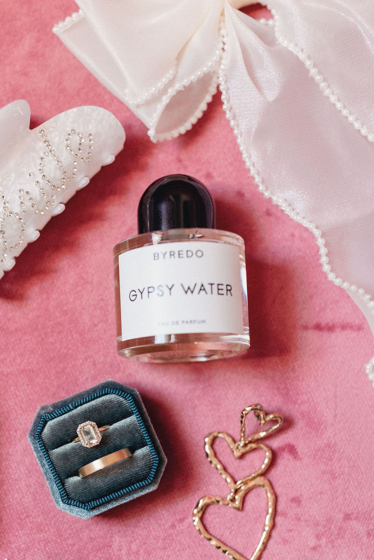 Luxury perfume bottles and engagement rings displayed on pink velvet with white lace ribbons and delicate gold jewelry.