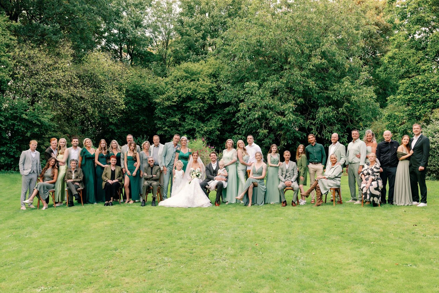 Wedding party members pose for group photos on a lush green lawn surrounded by trees.