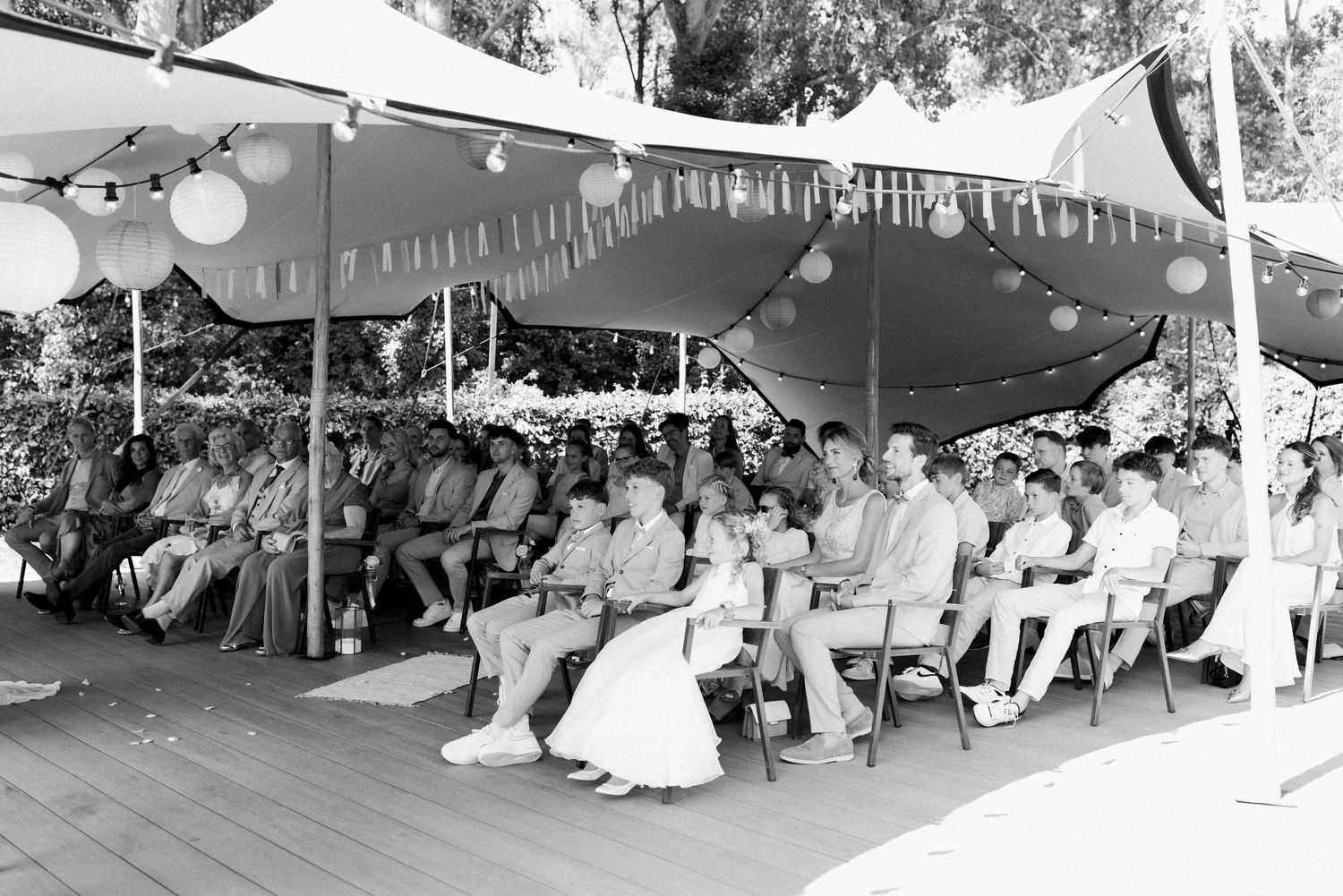 A panoramic view of people sitting under a tent at an outdoor gathering with hanging lights and decorations.