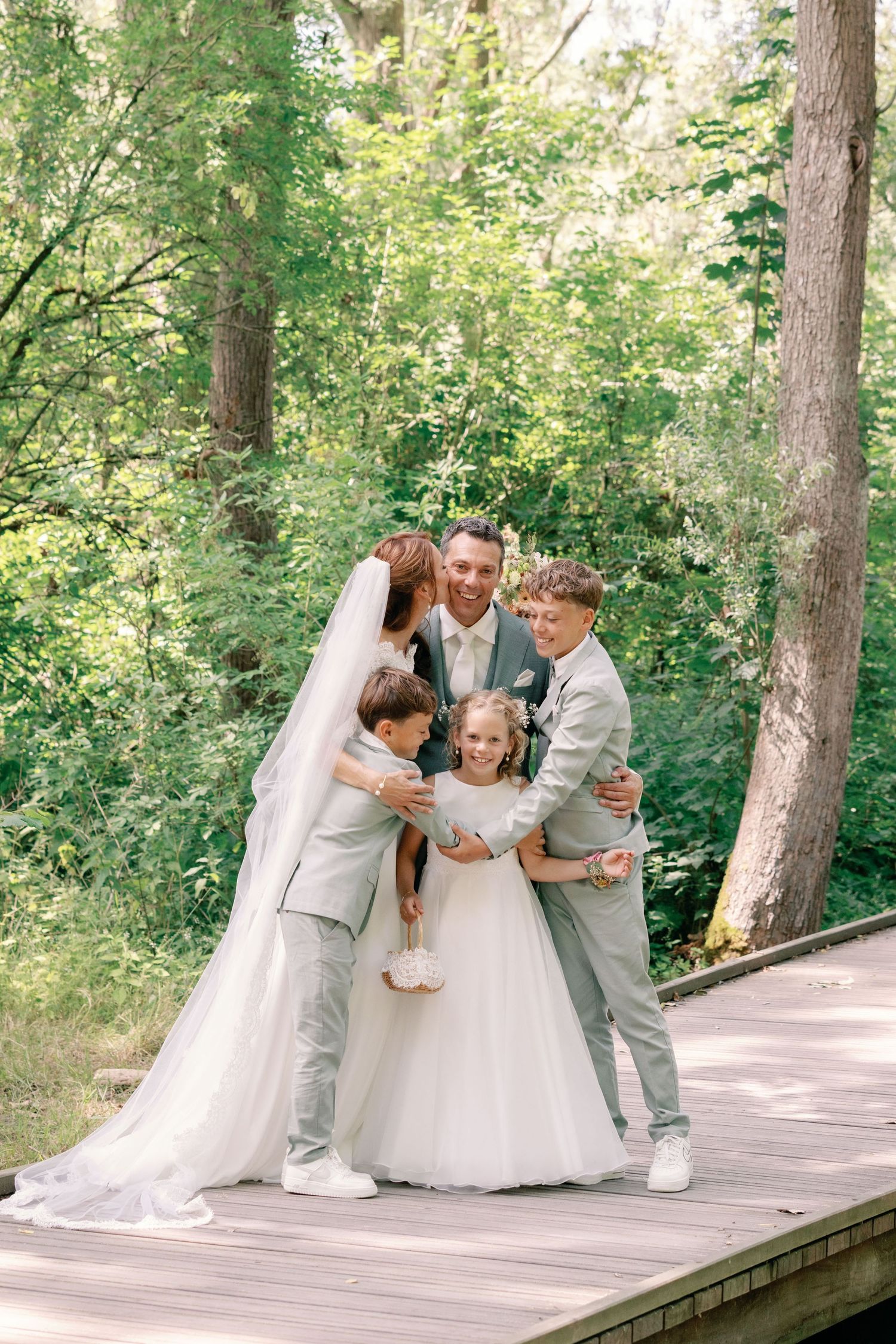 Wedding party shares a joyful moment on a wooden deck surrounded by lush green trees in a natural setting.