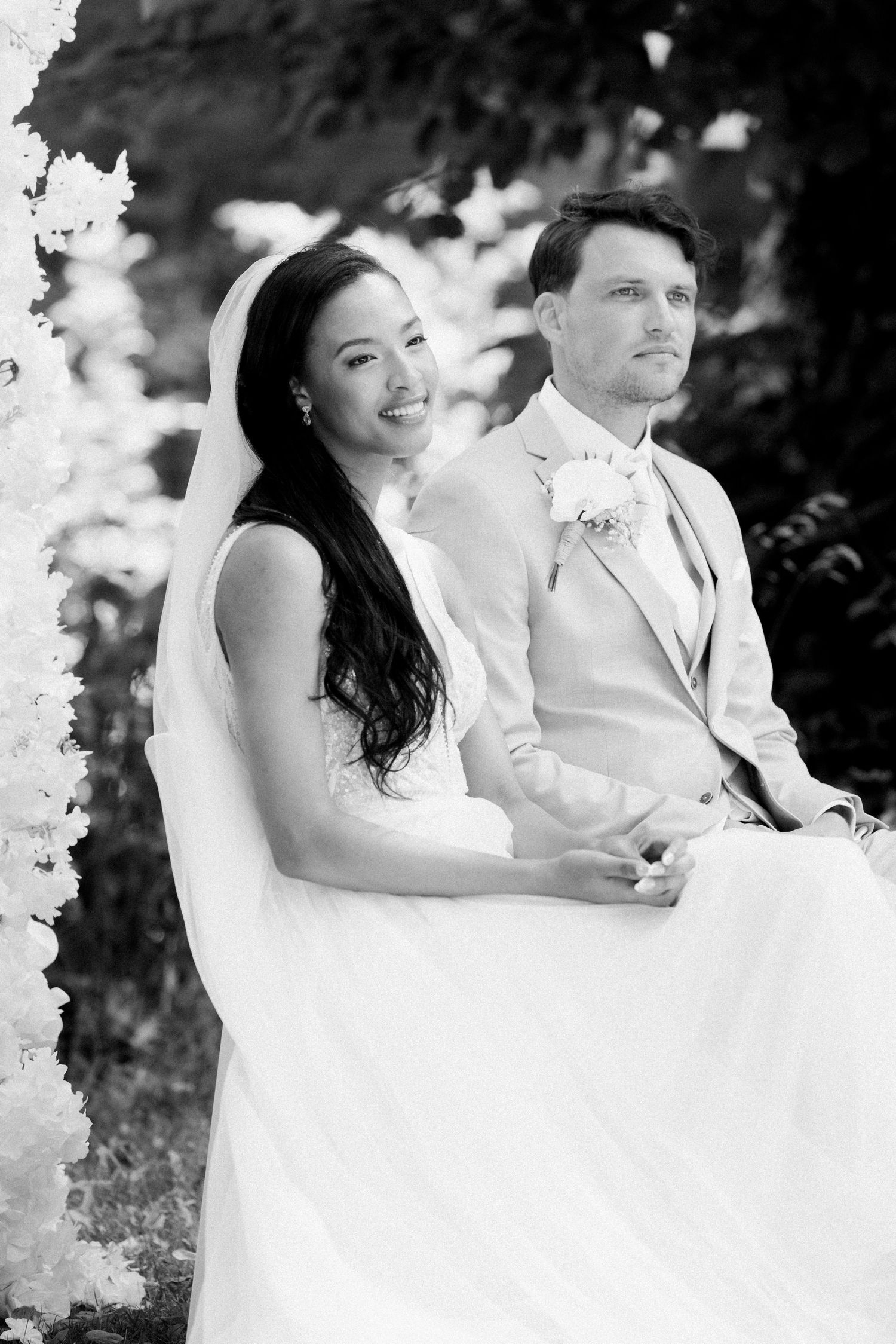 A bride and groom sit together outdoors in black and white wedding photos showing tender moments between them.