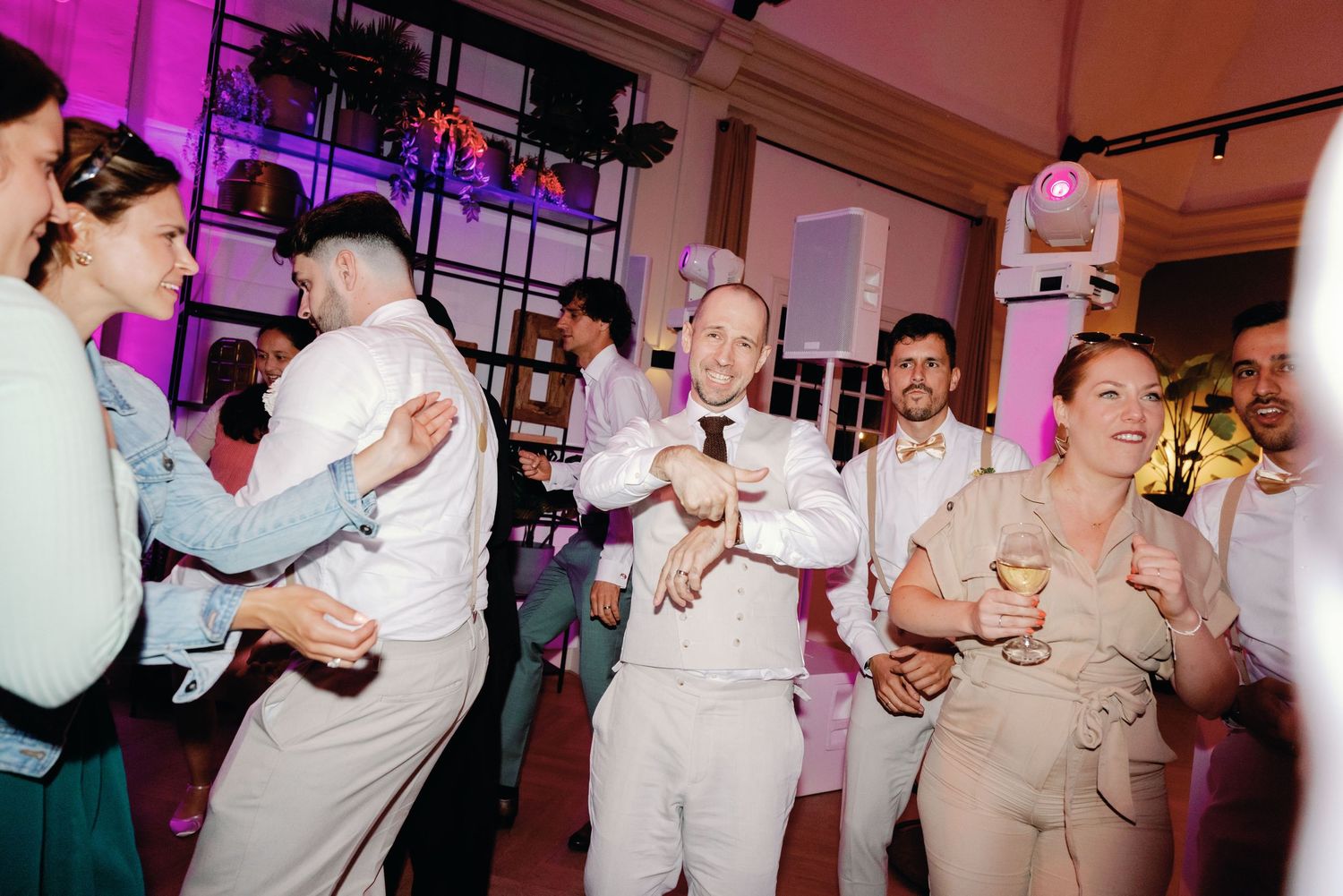 Wedding guests dance and celebrate on a lively dance floor illuminated by pink and purple lighting.